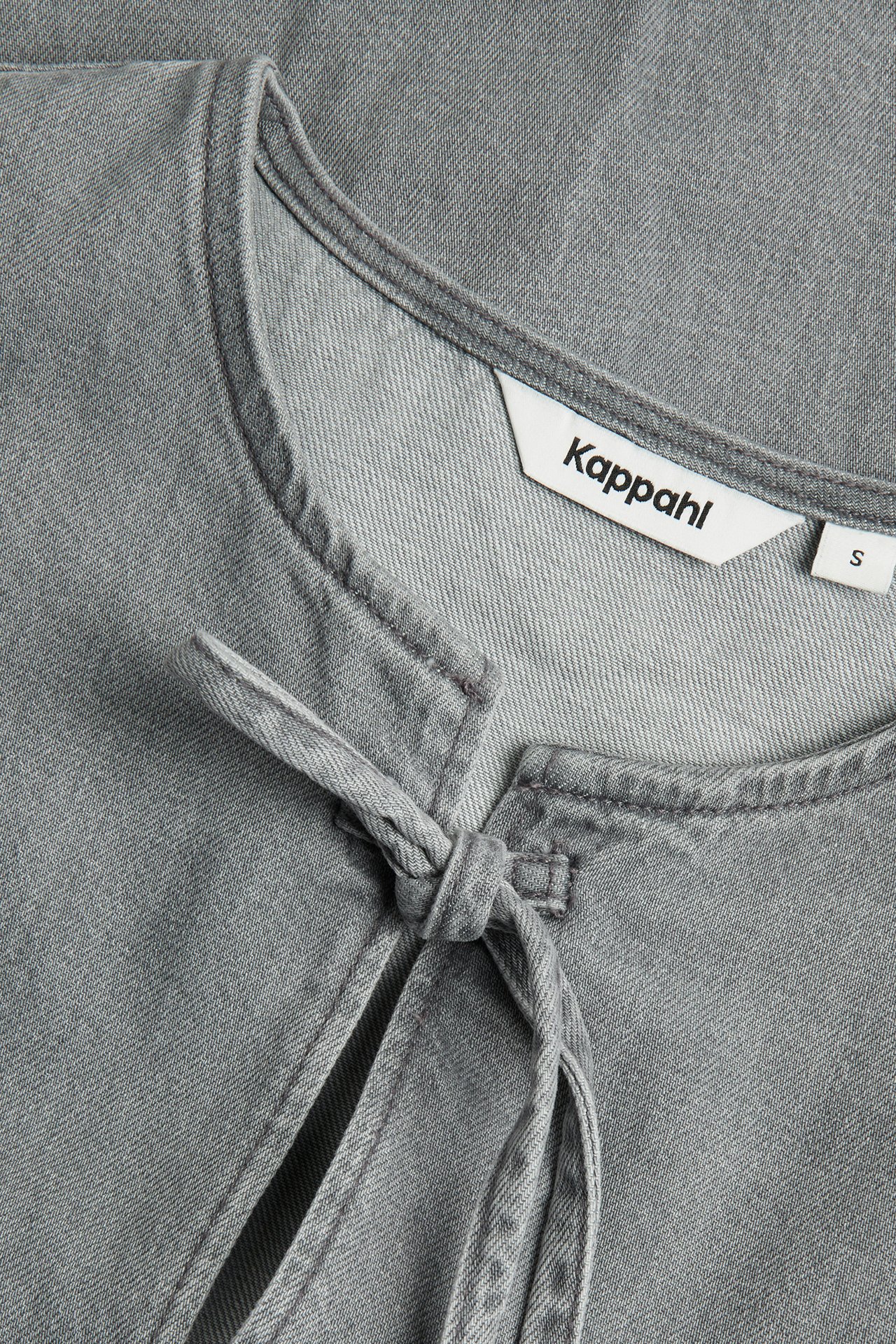 Närbild av en ljusgrå bomullsdenimblus med rund halsringning och knytband framtill.