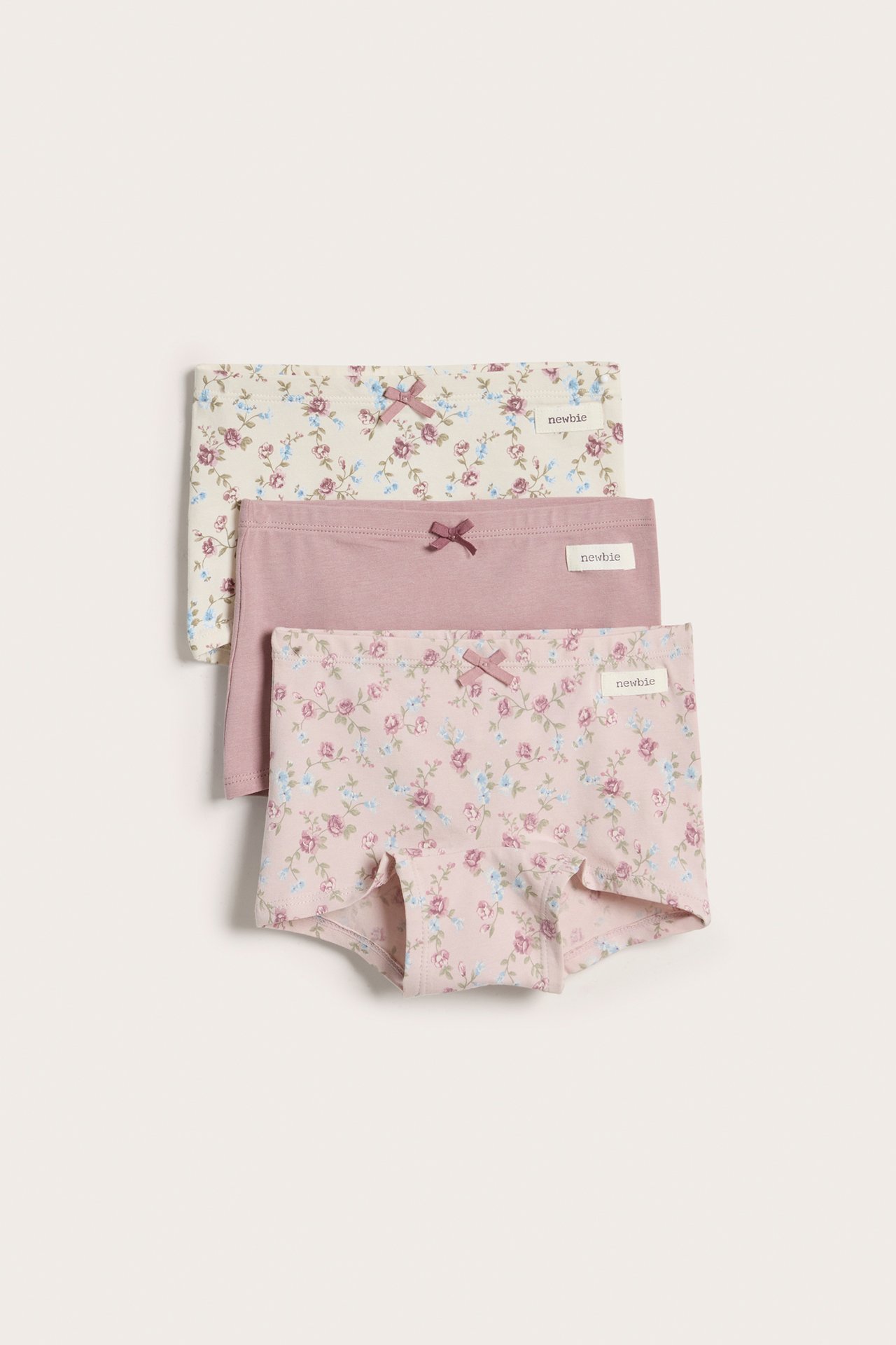 Frontvisning av tre stablede barne-hipstertruser i jersey: to blomstrete, en ensfarget rosa, alle med sløyfer.
