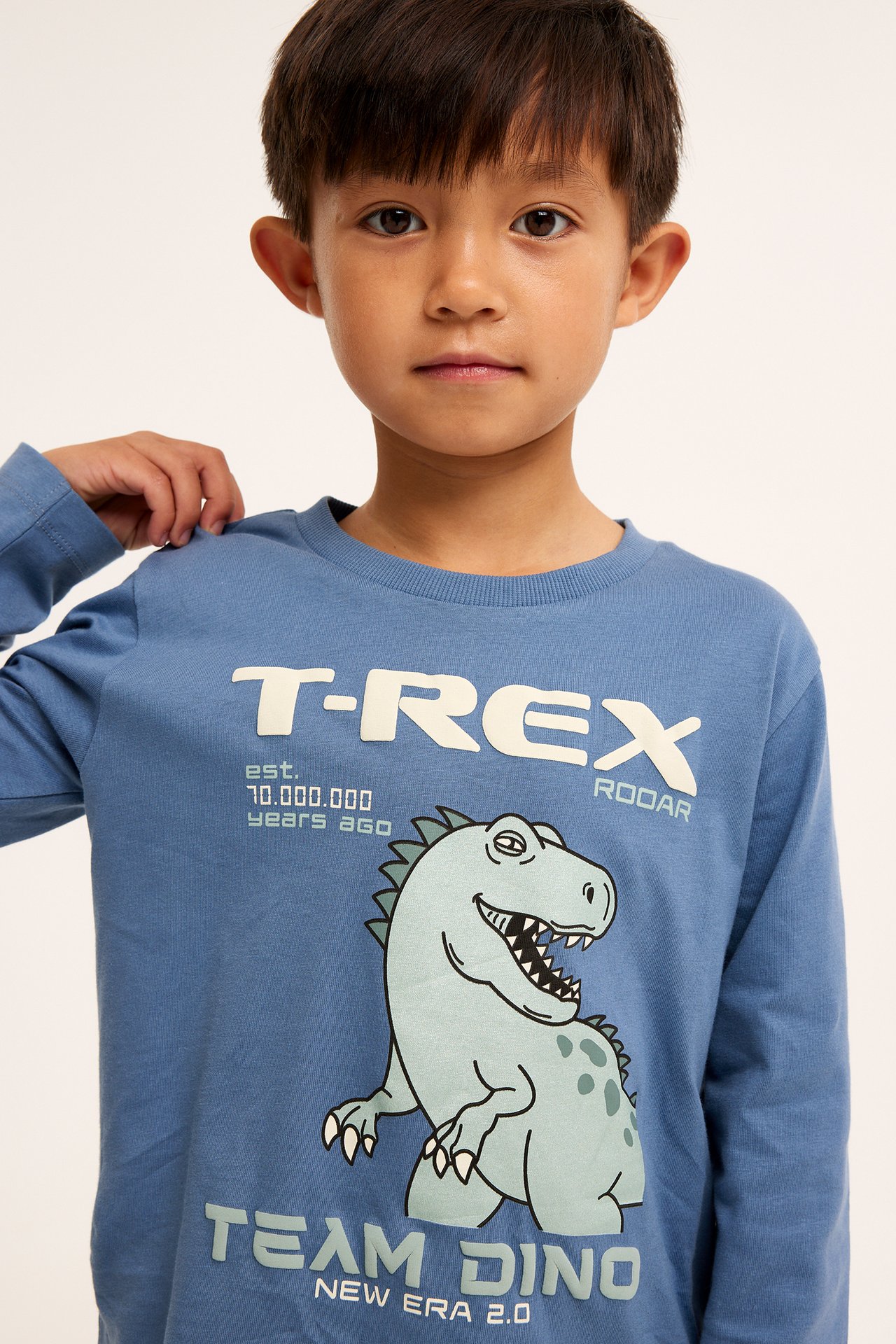 Edestä kuvattu lapsi sinisessä pitkähihaisessa puuvillapaidassa, jossa T-rex- ja tekstipainatus.