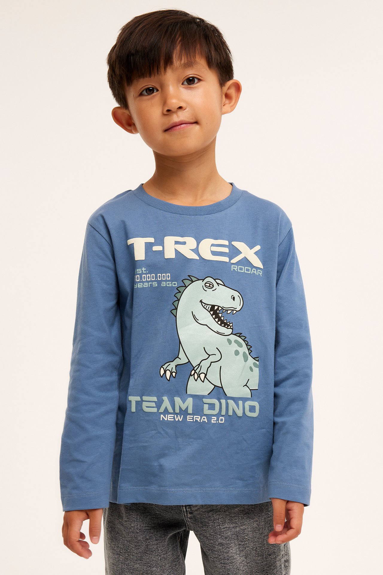 Edestäpäin, lapsi yllään sininen puuvillainen pitkähihainen T-paita T-Rex-painatuksella ja harmaat farkut.