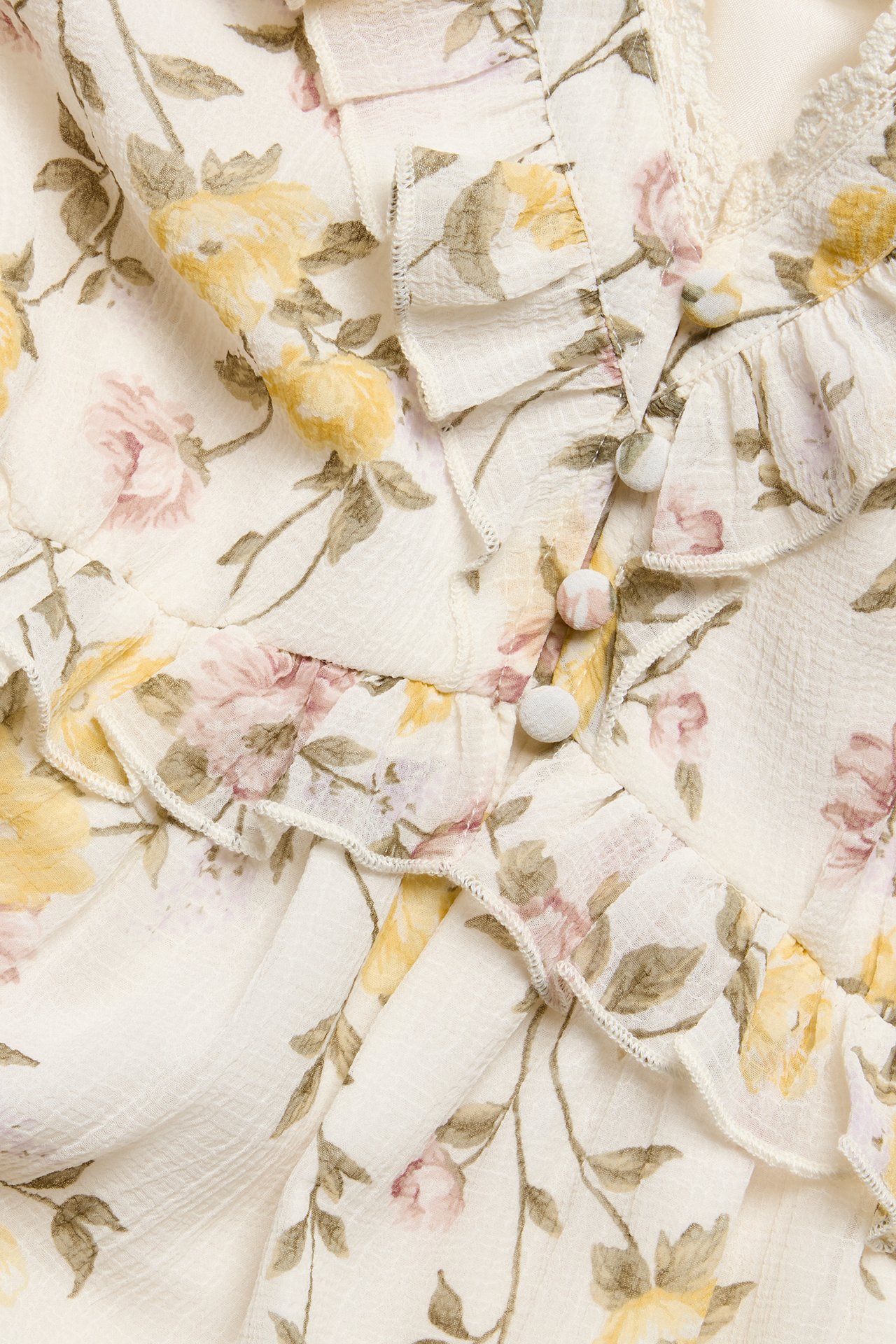 Nærbilde: Offwhite blomstret chiffongkjole med gule og rosa blomster, volanger, blonder og stofftrukne knapper.