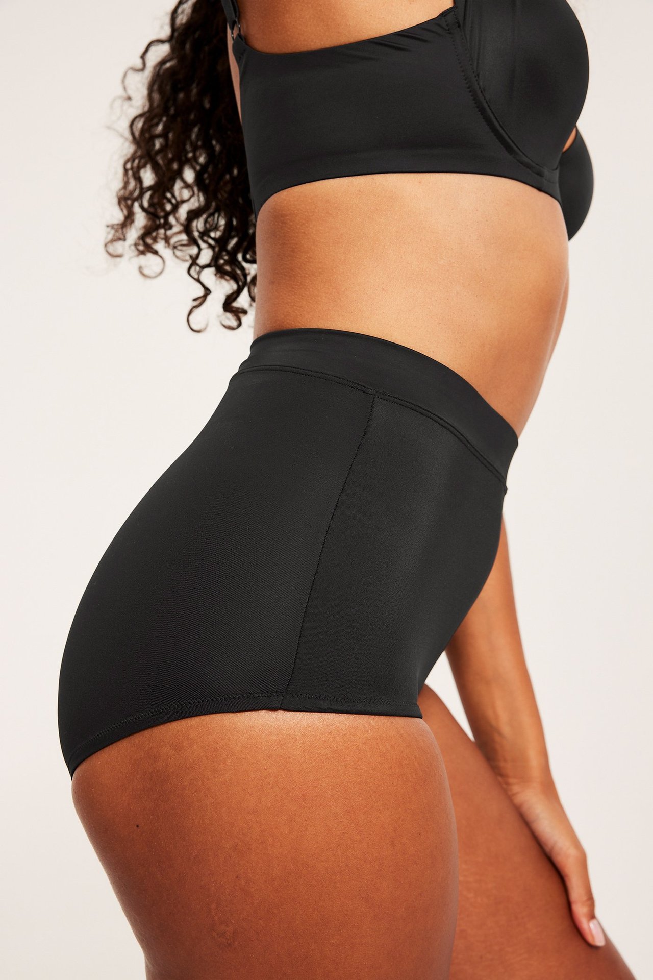 Naisen sivukuva mustissa korkeavyötäröisissä muotoilevissa hipster-alushousuissa ja mustissa rintaliiveissä.