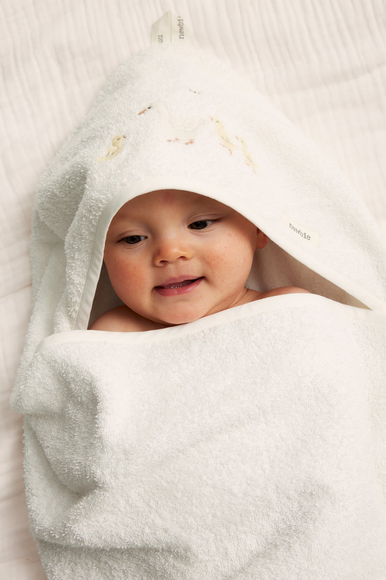 Närbild på en leende baby inlindad i en offwhite frottéhandduk med huva och ankbrodueri.
