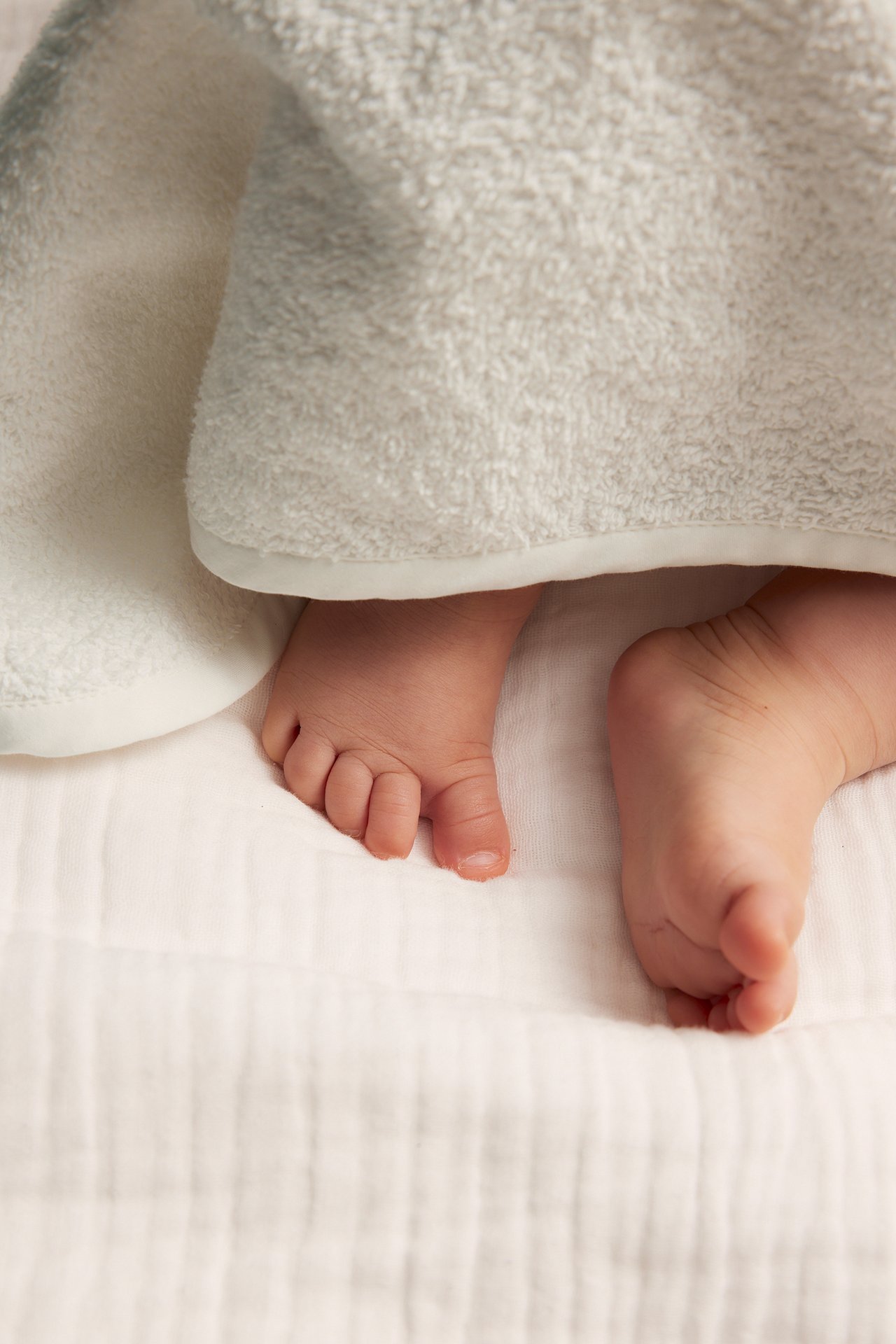 Närbild på en babys fötter som sticker fram under en mjuk, offwhite frottéhandduk.