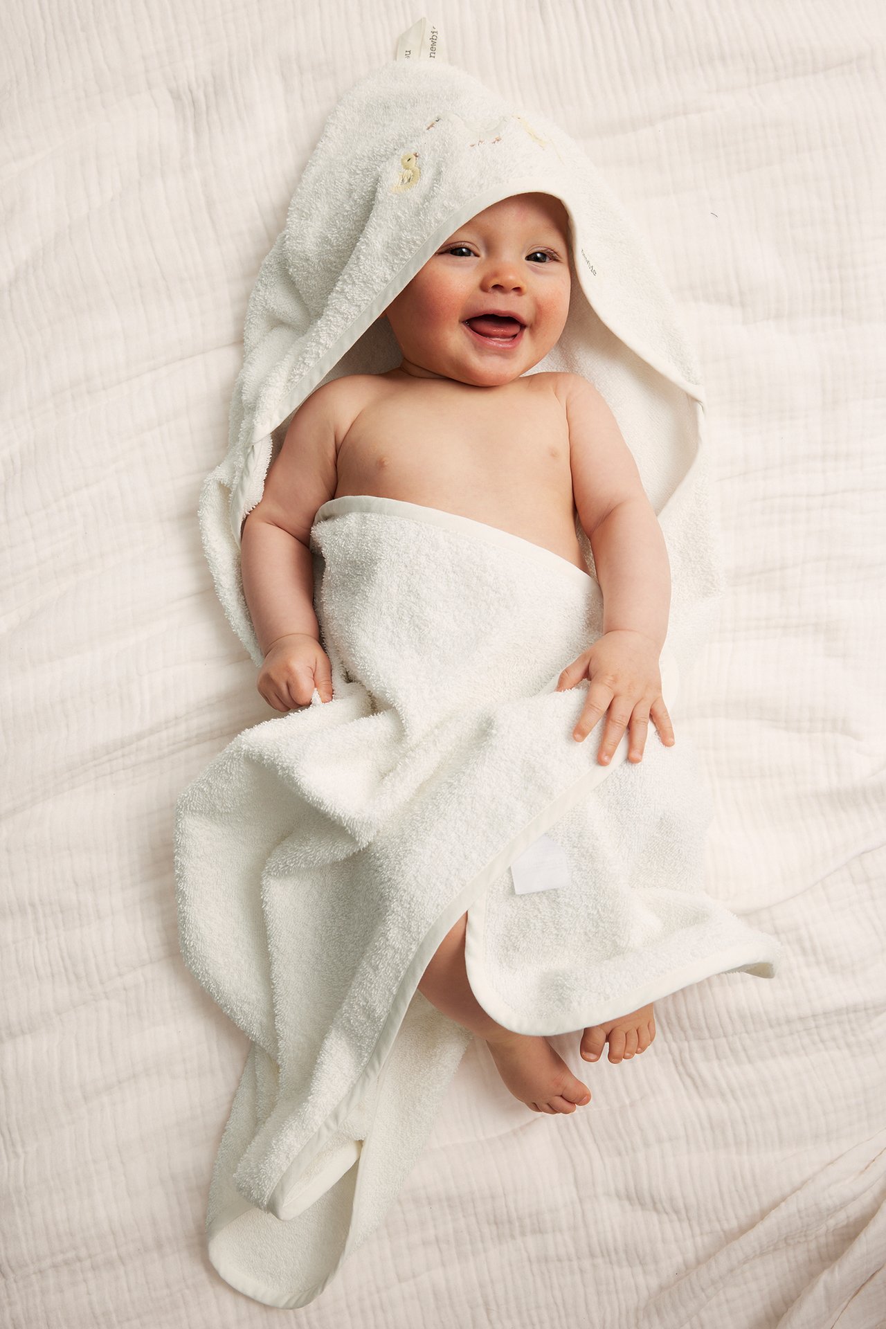 Uppifrån, leende baby inlindad i en naturvit frottéhandduk med huva och litet broderi.