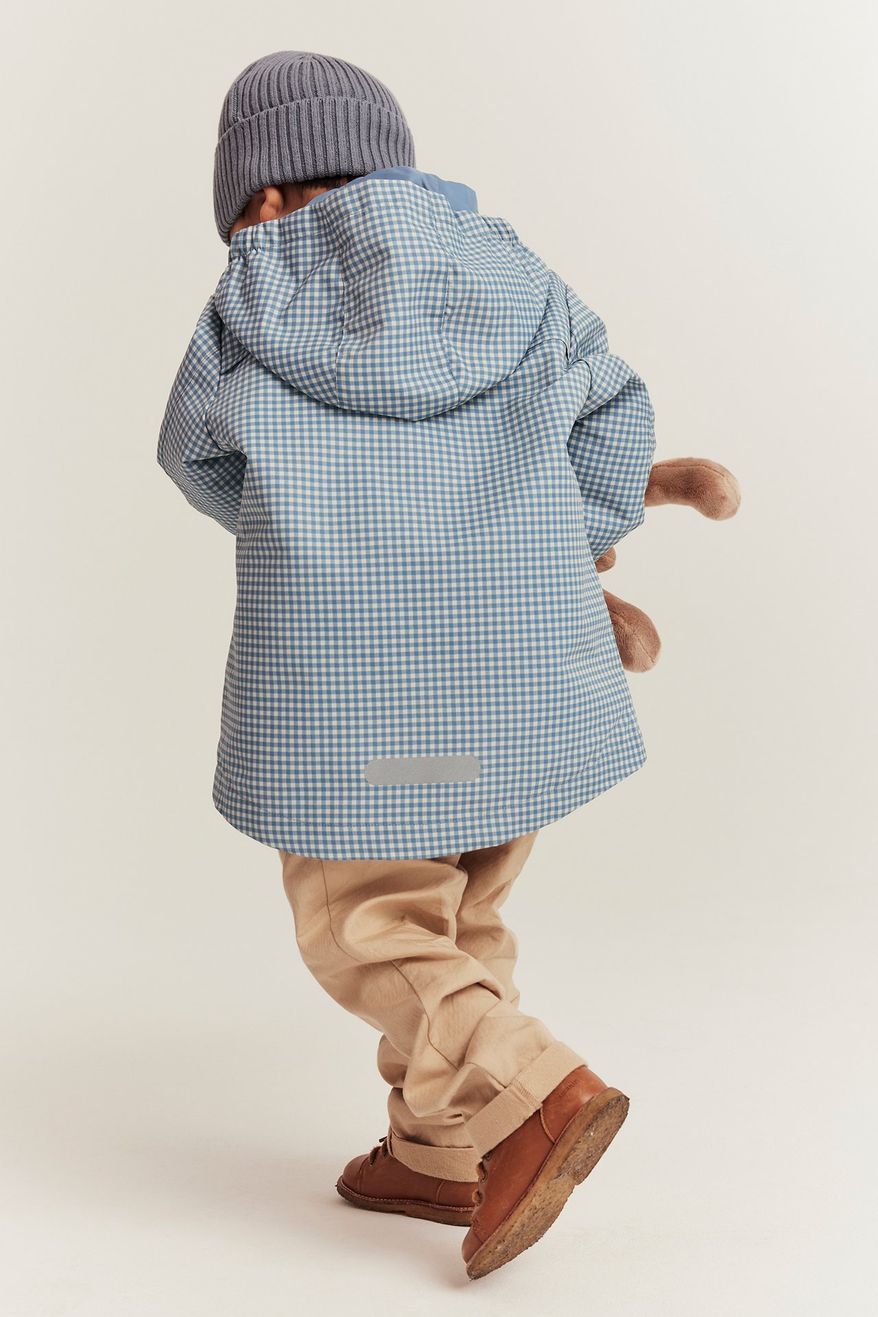 Widok z tyłu: dziecko w niebieskiej kurtce w kratę, szarej czapce, beżowych spodniach, brązowych butach, z pluszakiem.