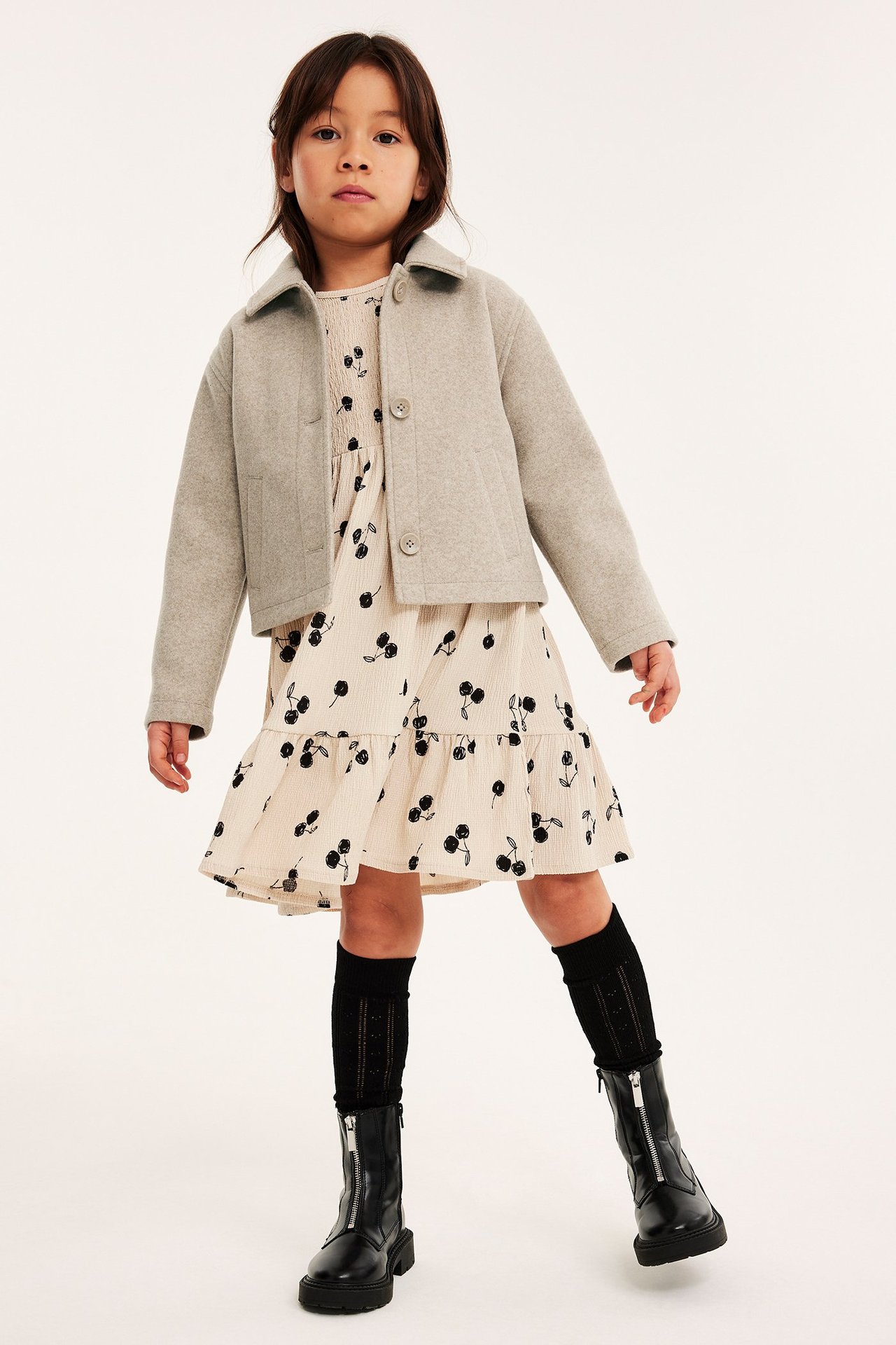 Edestä kuvattu lapsi yllään beigenvärinen lyhyt takki kirsikkakuvioisen mekon, mustien sukkien ja saappaiden päällä.