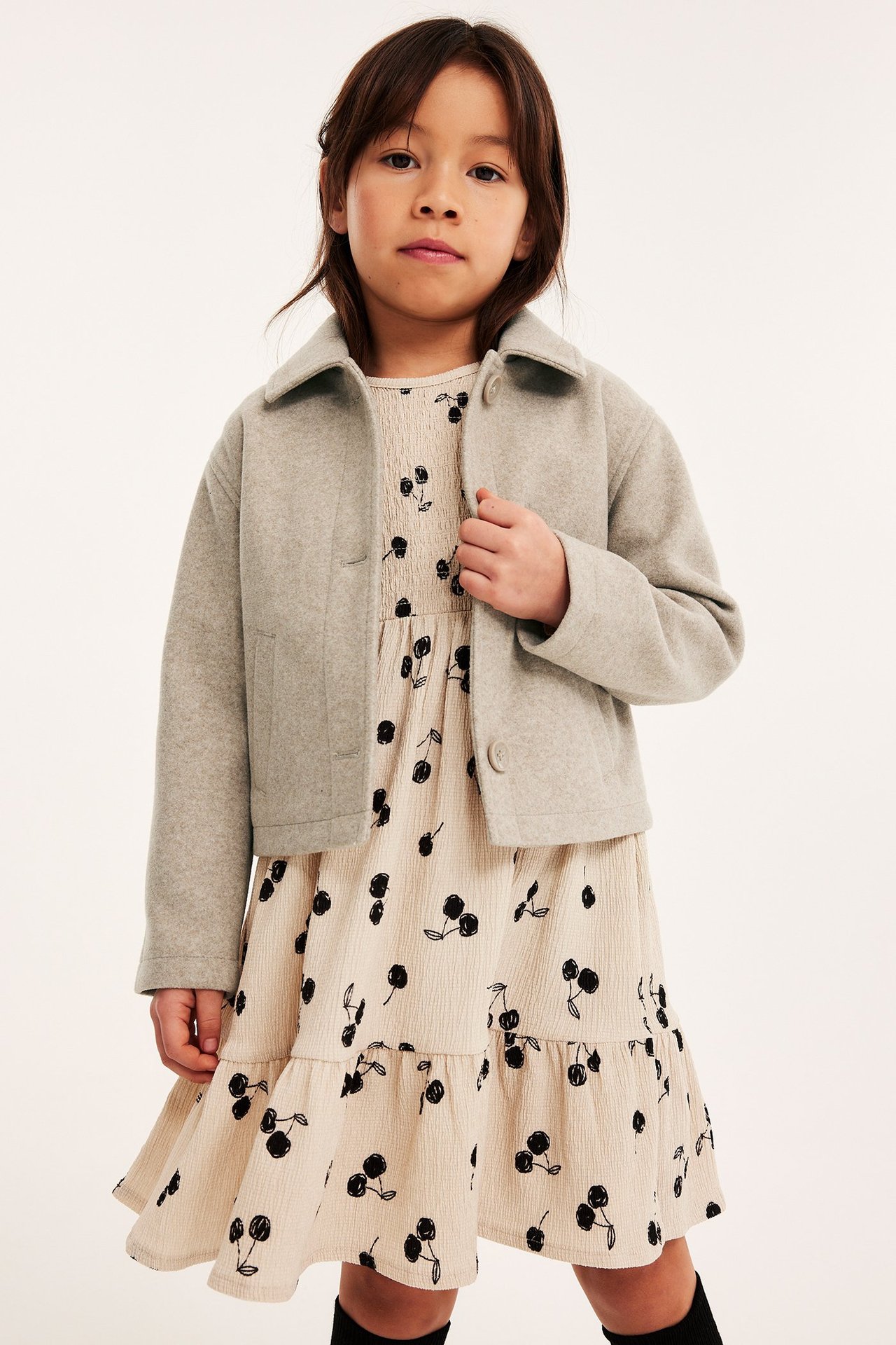 Edestä, lapsi beigessä meleeratussa lyhyessä takissa vaaleanbeigen kirsikkakuvioisen mekon päällä.