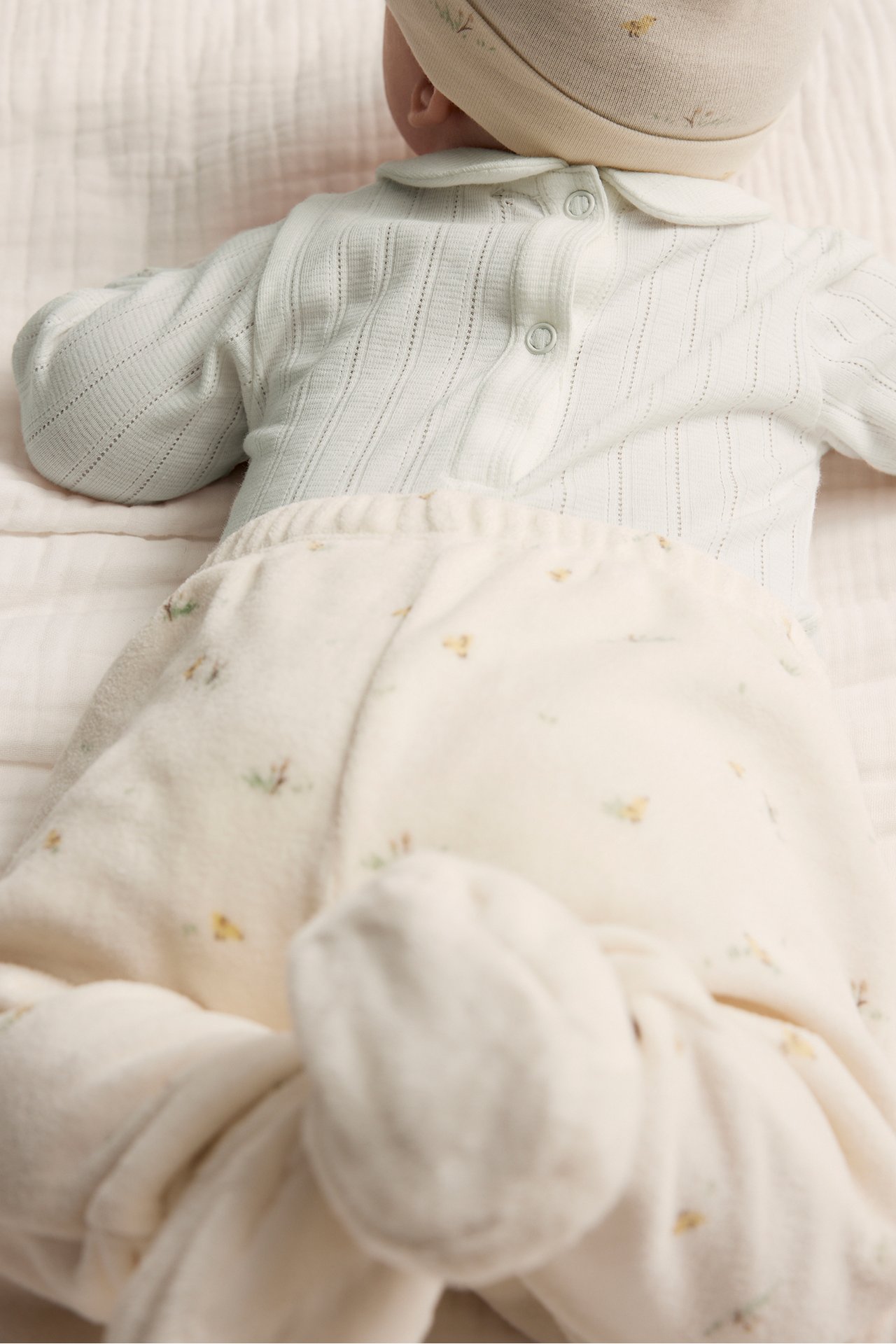 Niemowlę z tyłu: ecru body z kołnierzykiem, prążkowane, kwieciste spodnie i beżowa czapka.