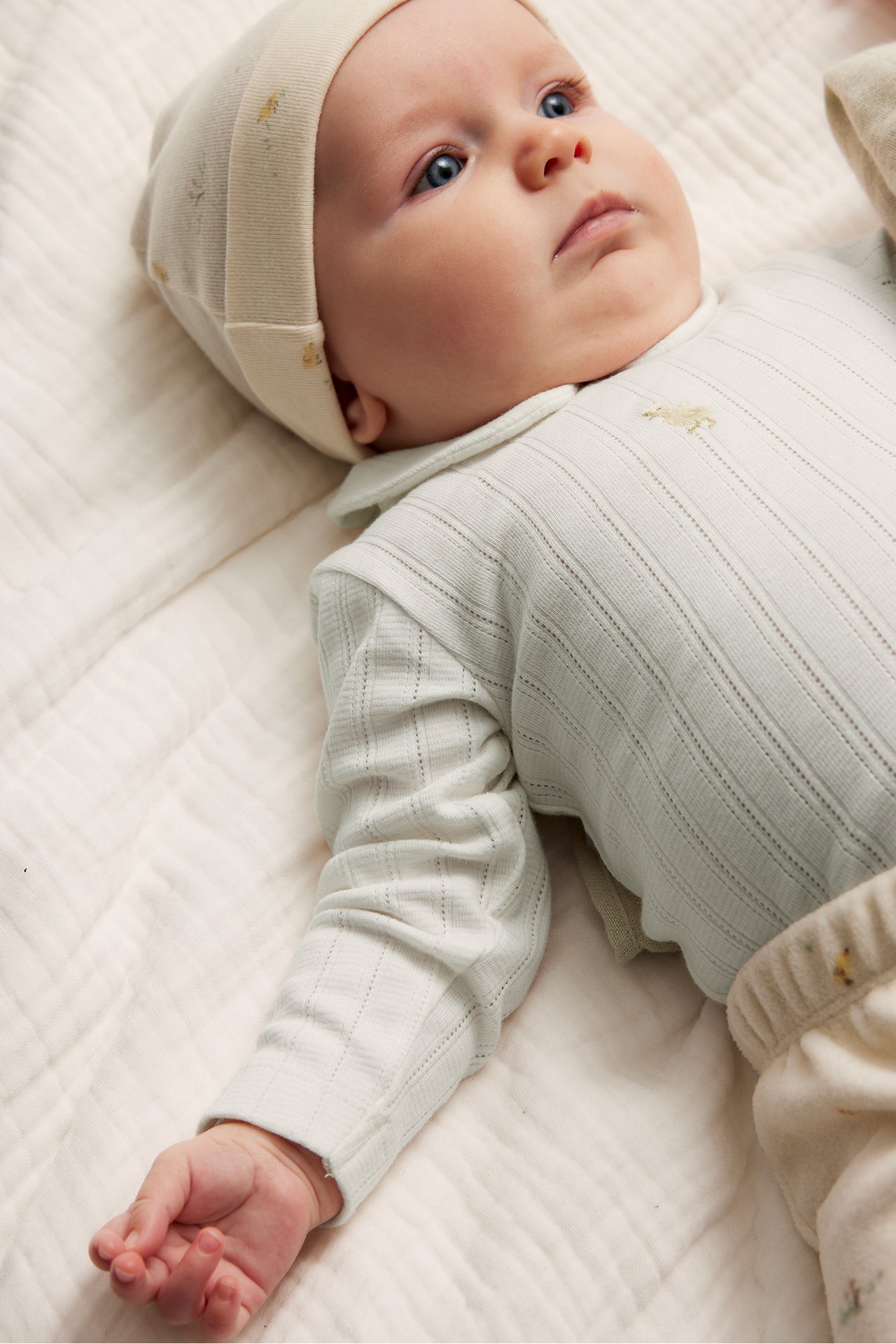 Zbliżenie dziecka w kremowym body pointelle z kołnierzykiem, haftem, pasującą czapką i spodniami.