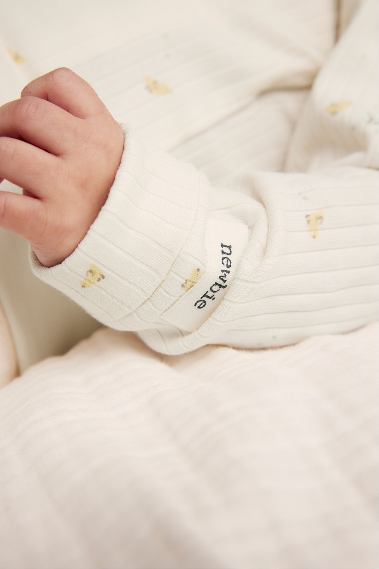 Nærbilde av babys arm i en offwhite ribbet bomullsbody med lite allovermønster.