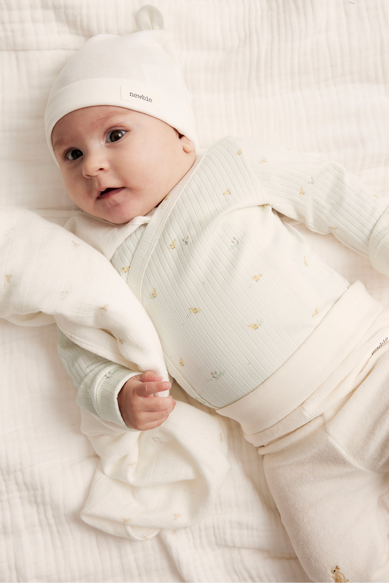 Ovenfra: Baby i offwhite ribbet omslagsbody med blomstermønster, offwhite lue, bukser, holder teppe.