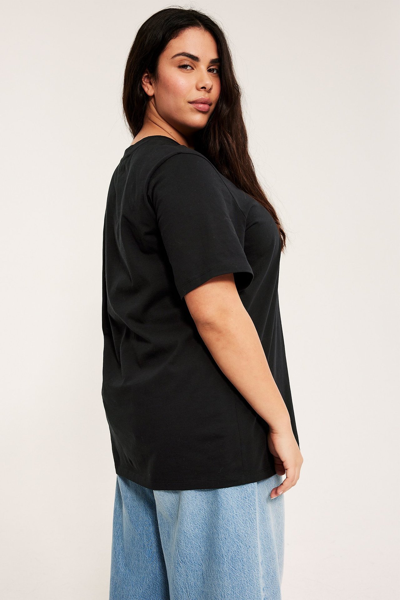 Sidevisning av kvinne i svart lang bomulls t-skjorte tunika med rund hals og lyseblå jeans.