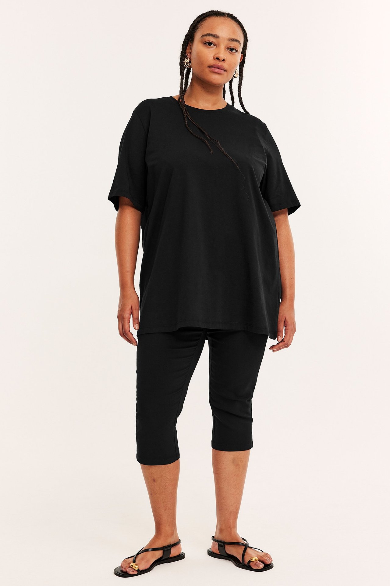 Framifrån: kvinna i lös svart bomulls lång t-shirt, svarta capribyxor och svarta platta sandaler.