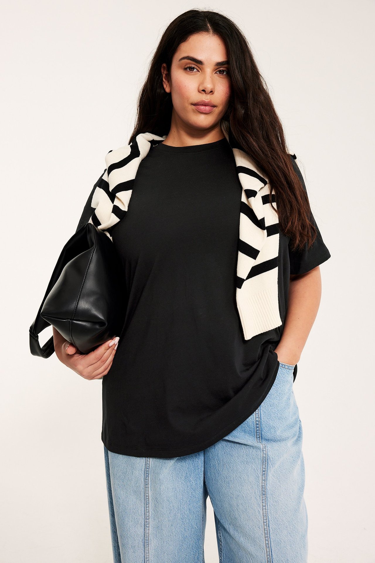 Frontutsikt: Kvinne med svart lang t-skjorte, lyseblå jeans, holder svart veske, stripete genser drapert.