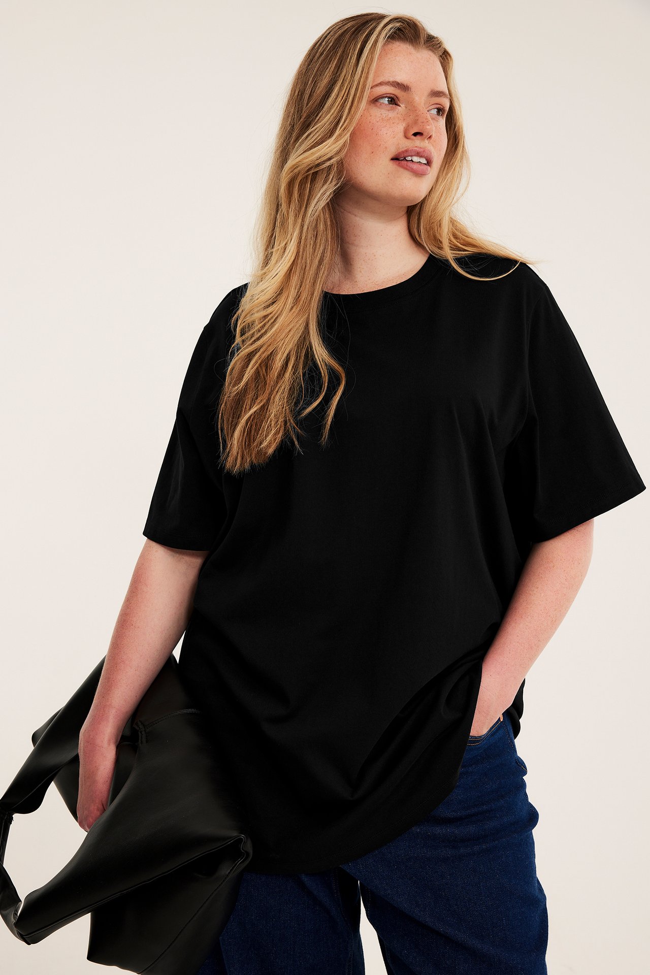 Frontbilde, kvinne i svart oversized t-skjorte, mørkeblå jeans, holder en svart veske.