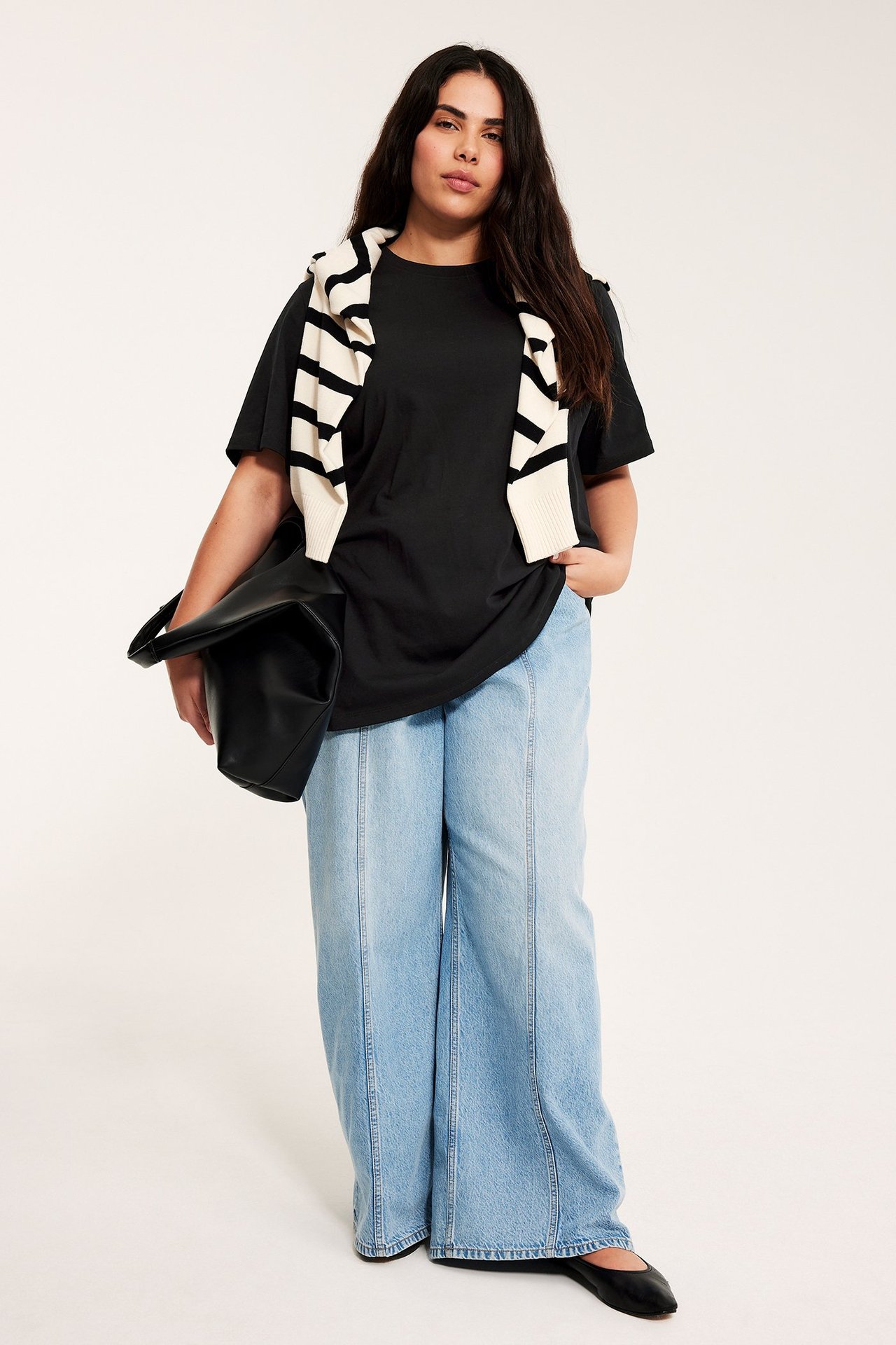 Front: Kvinne i svart lang t-skjorte, lyseblå jeans, svarte sko. Holder svart veske, stripet genser over skuldrene.