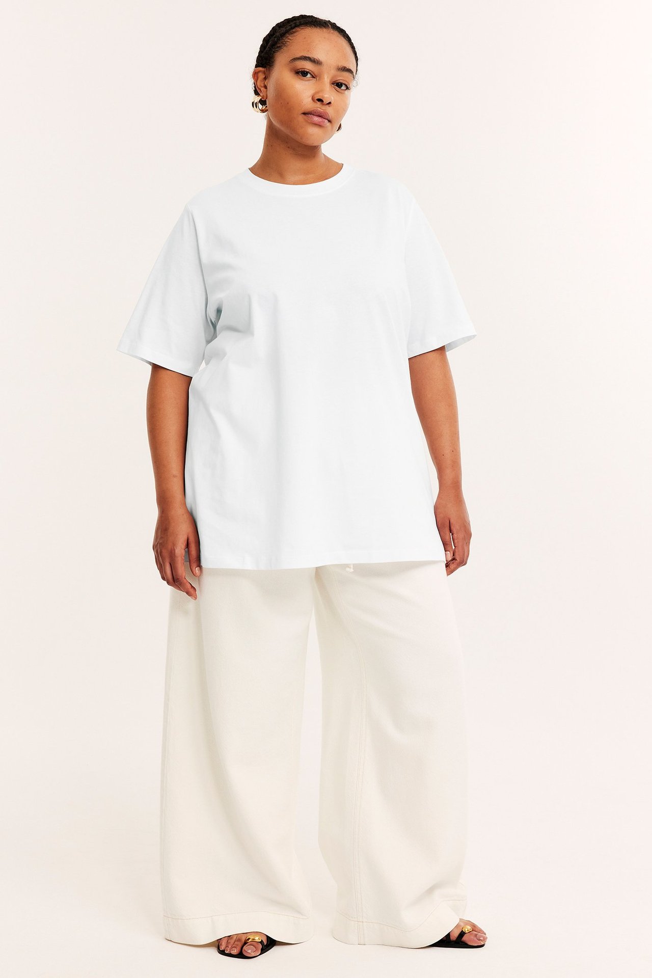 Forfra: kvinne i løs hvit lang bomulls-t-skjorte, hvite vide bukser, svarte sandaler og gulløreringer.