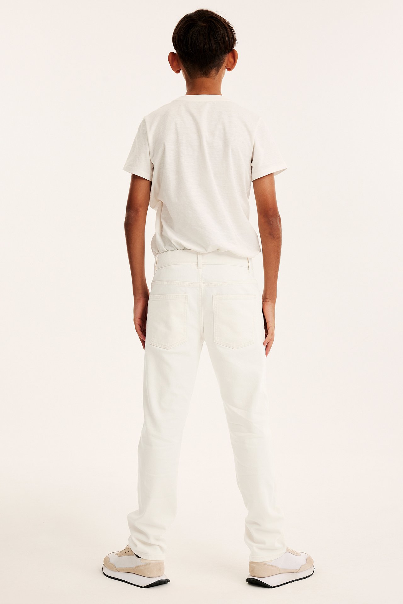 Widok z tyłu: Dziecko w off-white prostych jeansach z wysokim stanem, jasnym T-shircie i beżowych sneakersach.
