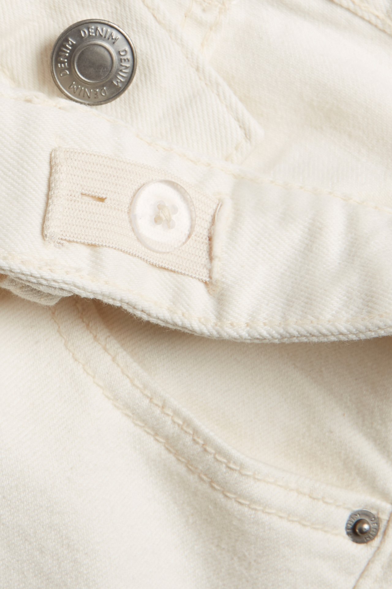 Zbliżenie na dziecięce off-white proste jeansy denimowe, z metalowym guzikiem i wewnętrzną regulowaną gumką.