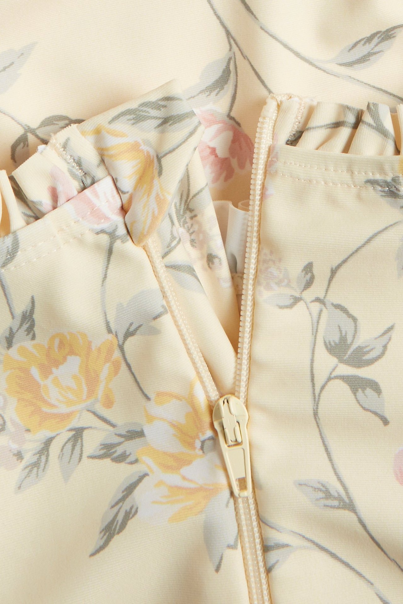 Lähikuva vaaleankeltaisesta kukkakuosisesta UV-uimapuvusta, jossa on röyhelöitä ja vetoketju.