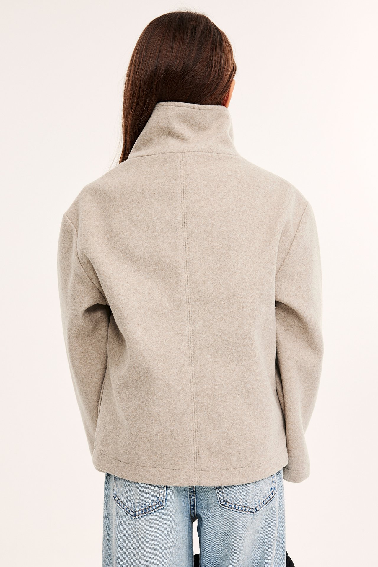 Bakifrån: ett barn i en kort, boxig melerad beige jacka med hög krage över ljusblå jeans.