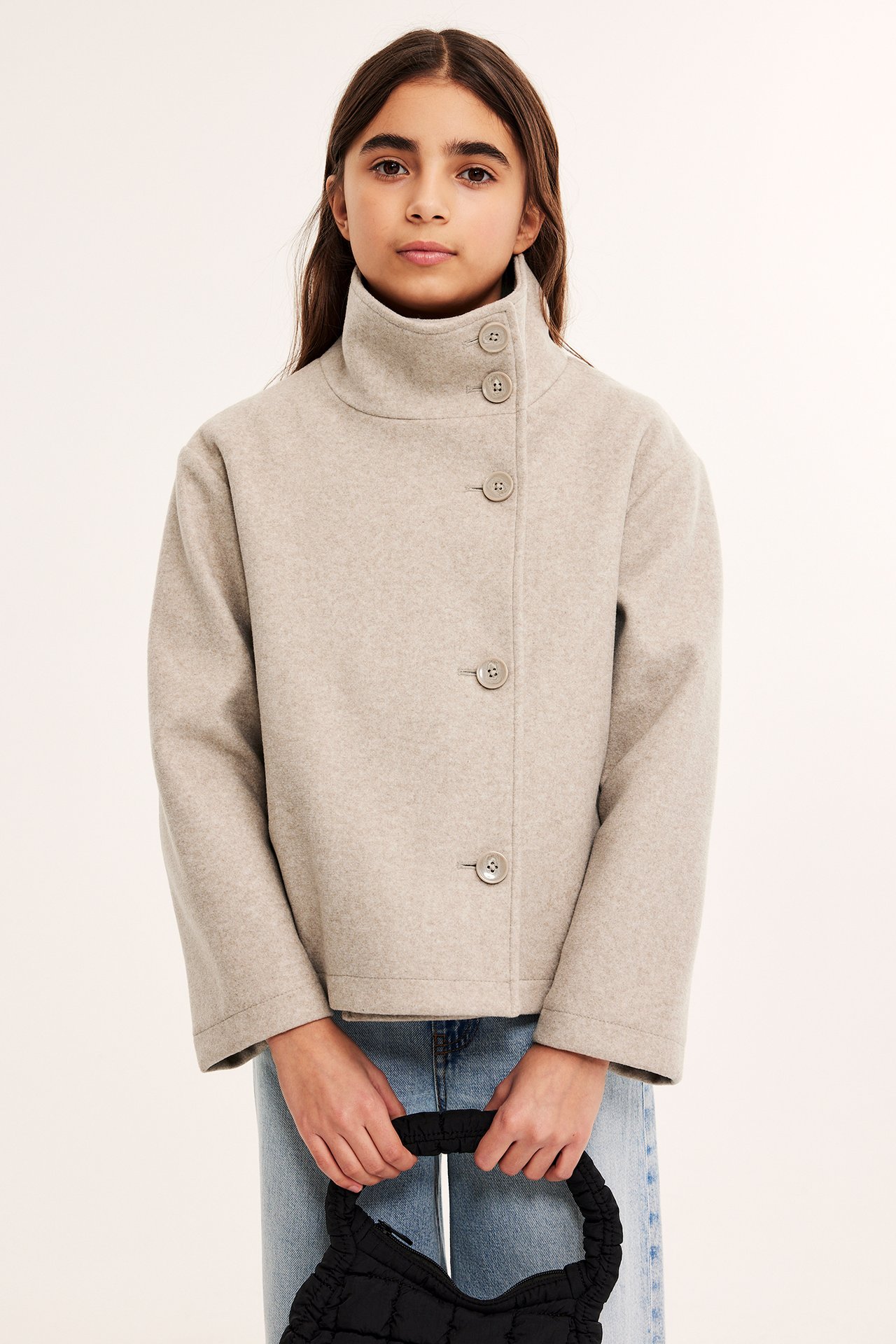 Framifrån: Barn bär en beige melerad kappa med hög krage, knappar, över jeans, håller en svart väska.