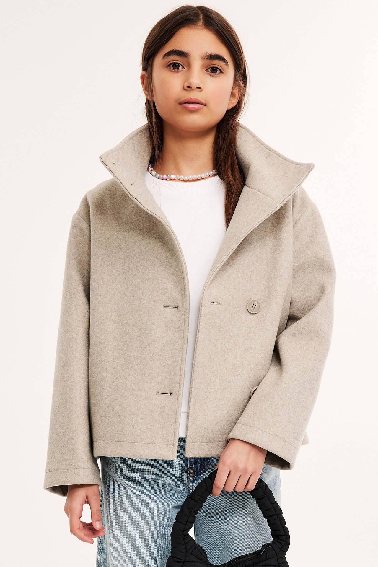 Barn bär kort beige melerad jacka, hög krage, vit t-shirt, jeans, håller svart quiltad väska.