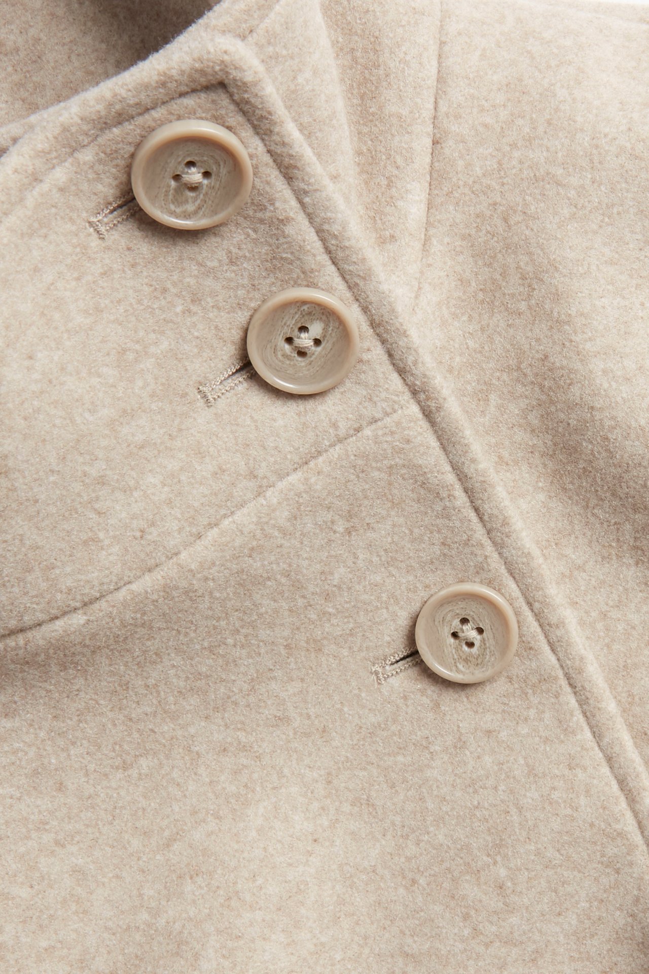 Närbild av en melerad beige kort kappa med tre ljusbeige knappar och knapphål.