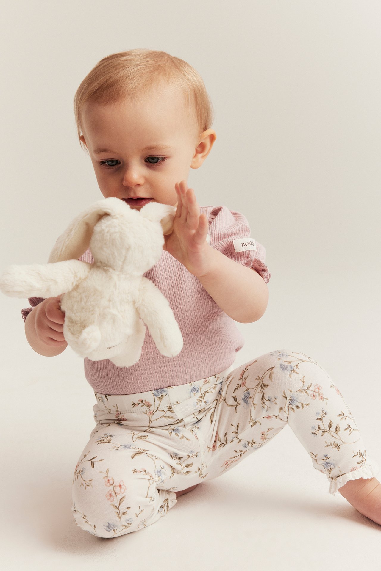 Frontvy: Baby i rosa ribbad body med puffärm, vita blommiga leggings, håller en vit fluffig kanin.