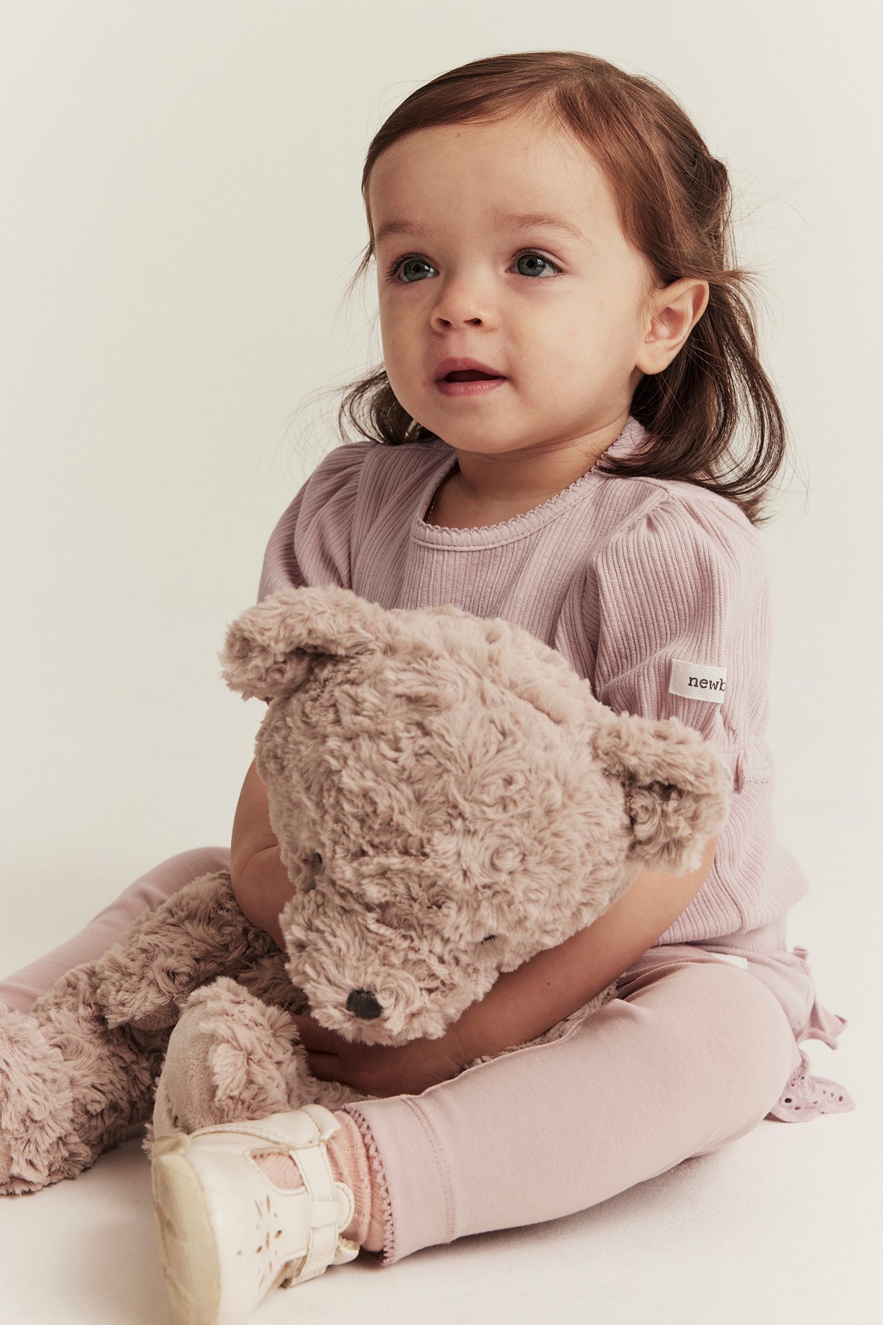 Framifrån, baby i rosa ribbad body med puffärm och rosa leggings, håller en brun nallebjörn.