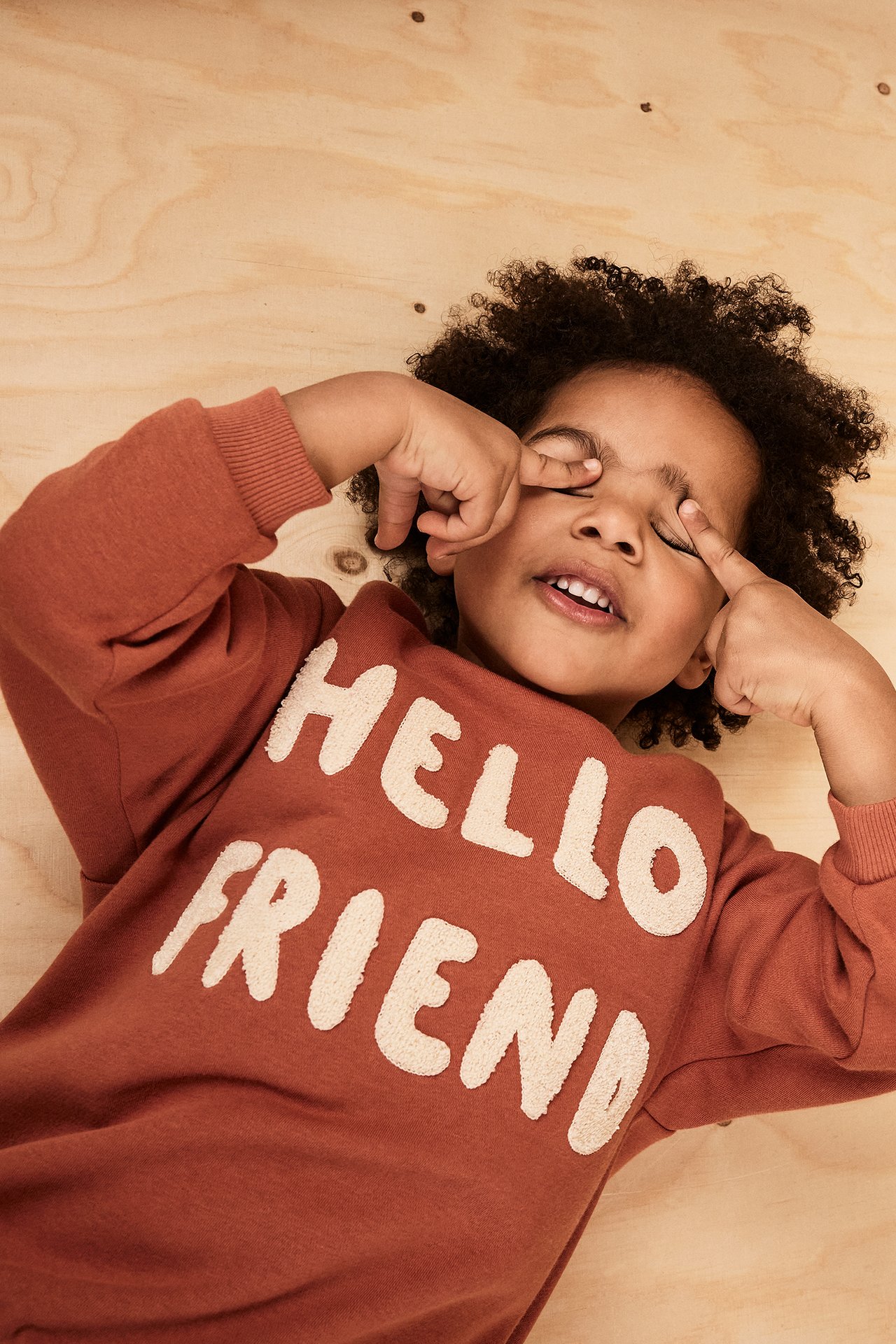 Nærbilde av et barn i en brun sweatshirt med hvit "HELLO FRIEND" frottéapplikasjon, gjør en morsom grimase.