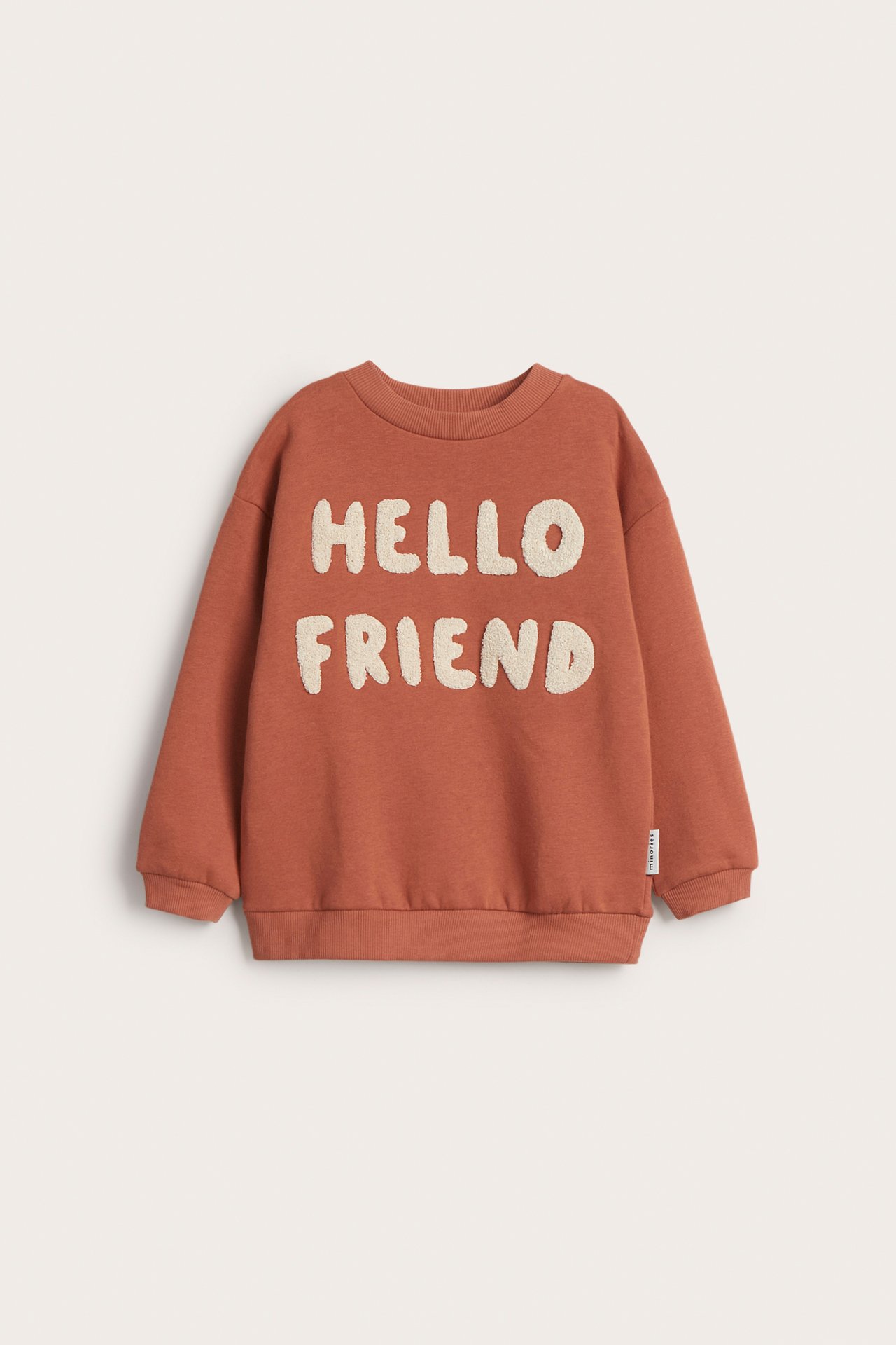 Frontvisning av en brun barnegenser med kremfarget 'HELLO FRIEND' frottéapplikasjon, ribbestrikkede mansjetter og kant.
