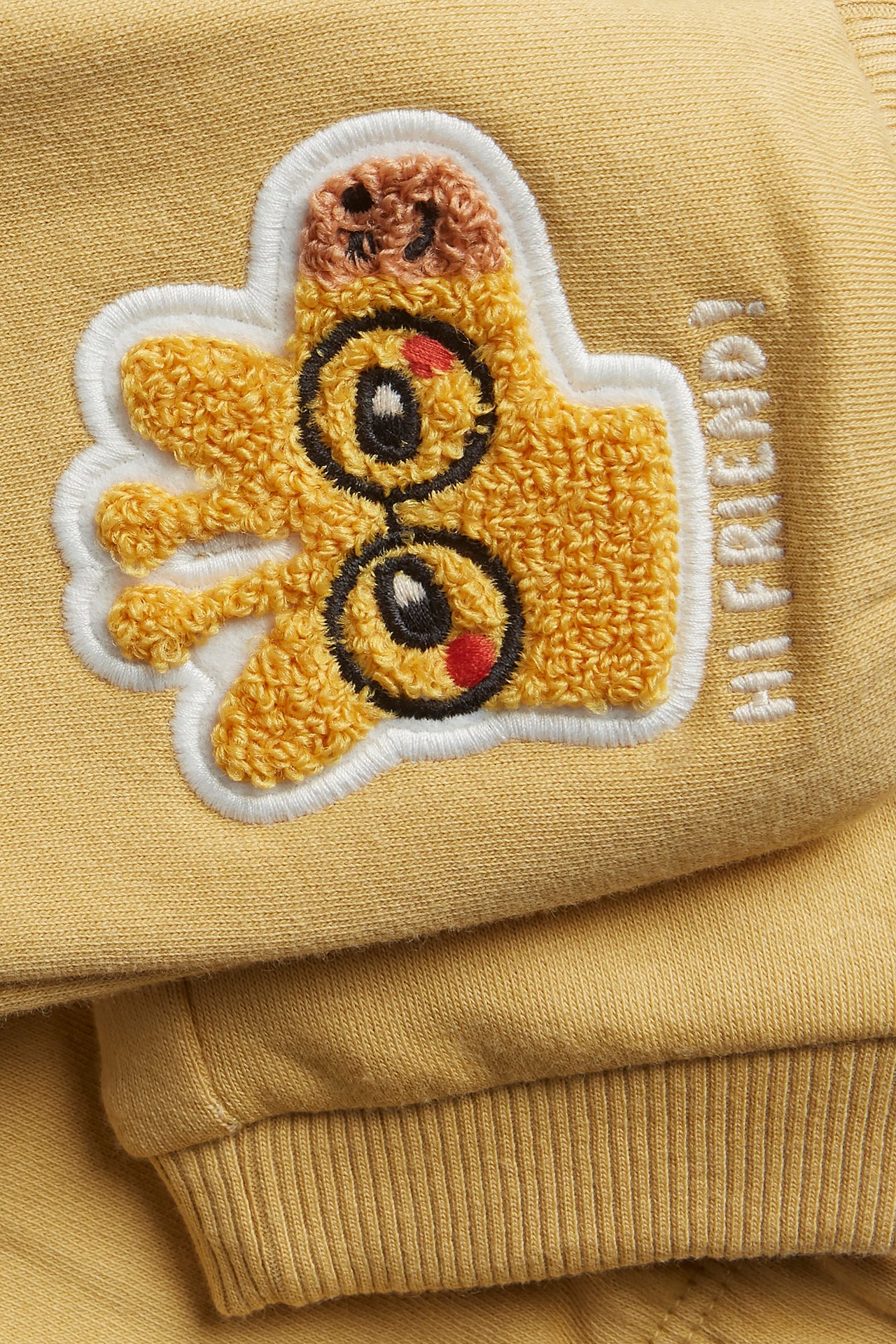 Närbild på gula babyjoggingbyxor med frottéapplikation av tumme upp och broderad text "HI FRIEND!".