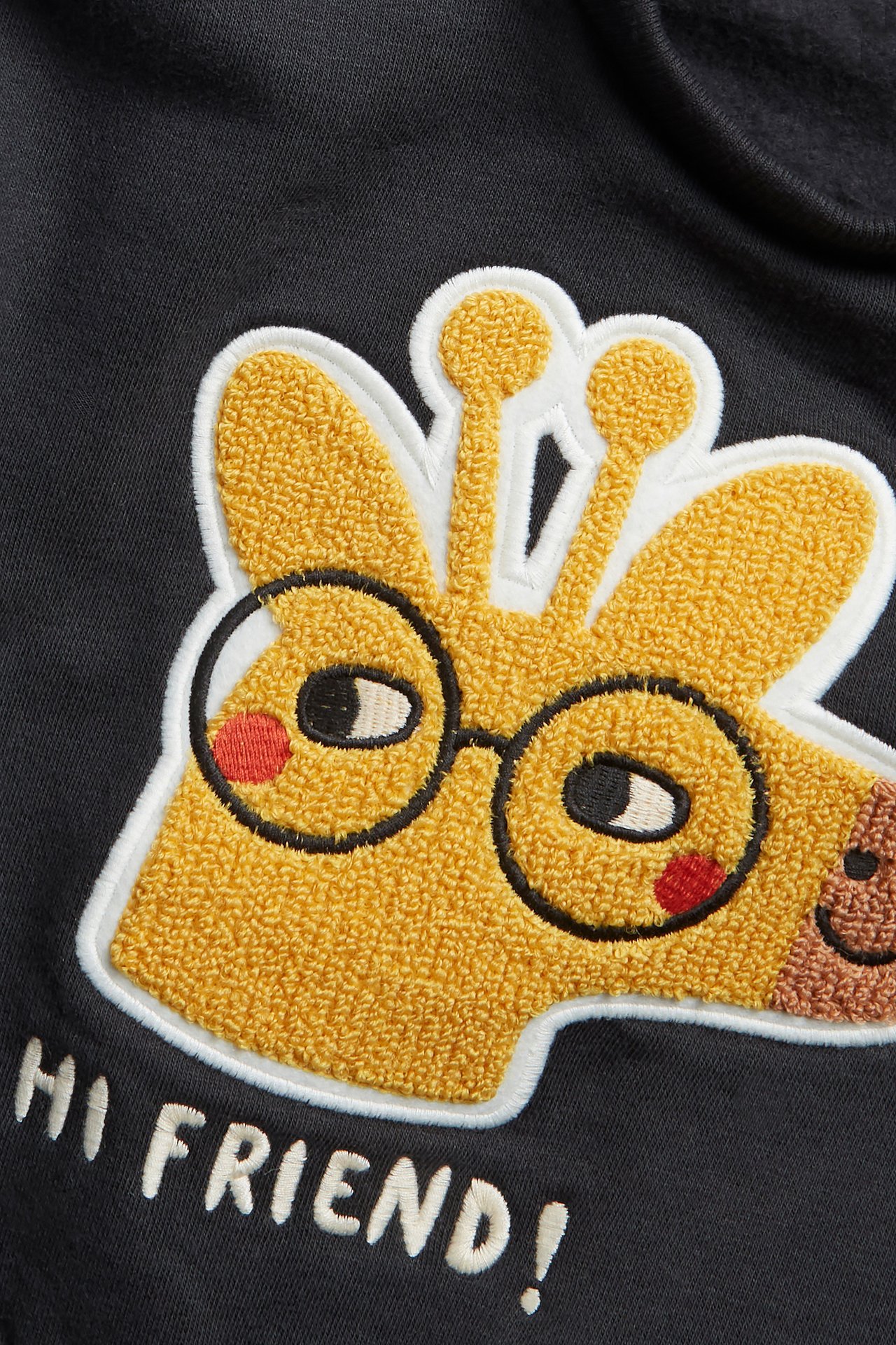 Nærbilde av svart babygenser med gul terryapplikasjon av sjiraff med briller og teksten 'HI FRIEND!'.