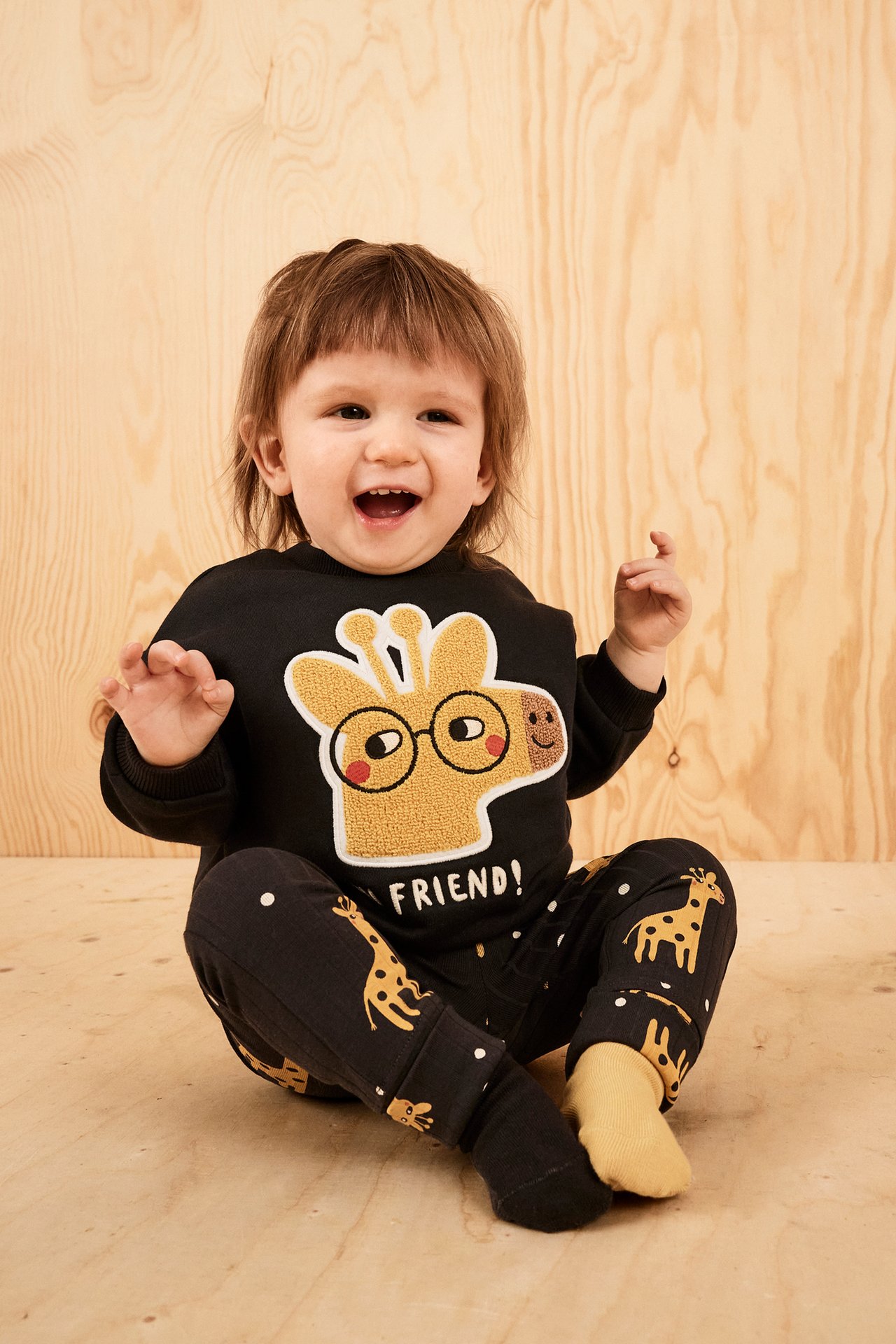 Forfra, smilende baby i svart sweatshirt med sjiraffapplikasjon, svarte mønstrede bukser og gule sokker.