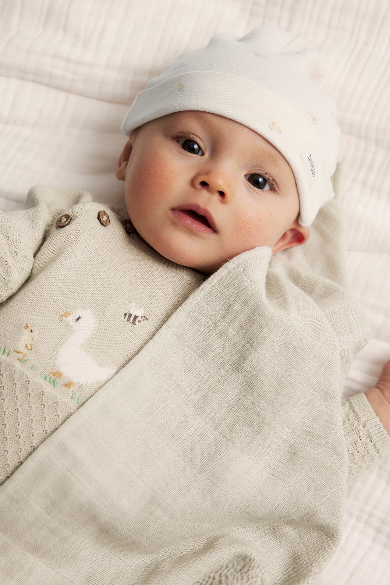 Närbild på baby i offwhite bomullsmössa med små mönster, stickad tröja med ankmotiv och ljusgrön muslinfilt.