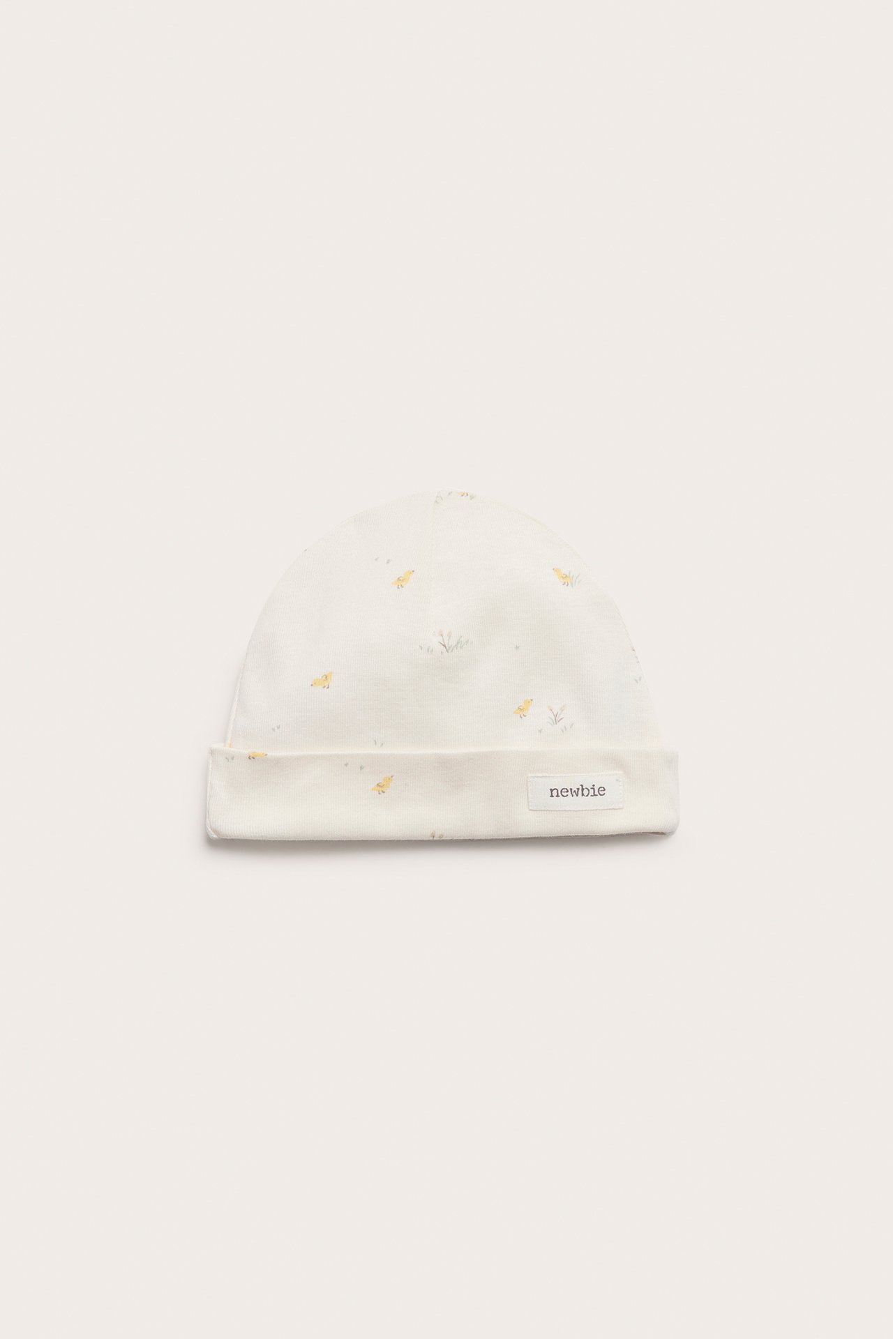 Frontvy av en offwhite bomullsbeanie för baby med litet kycklingmönster och 'Newbie'-etikett.