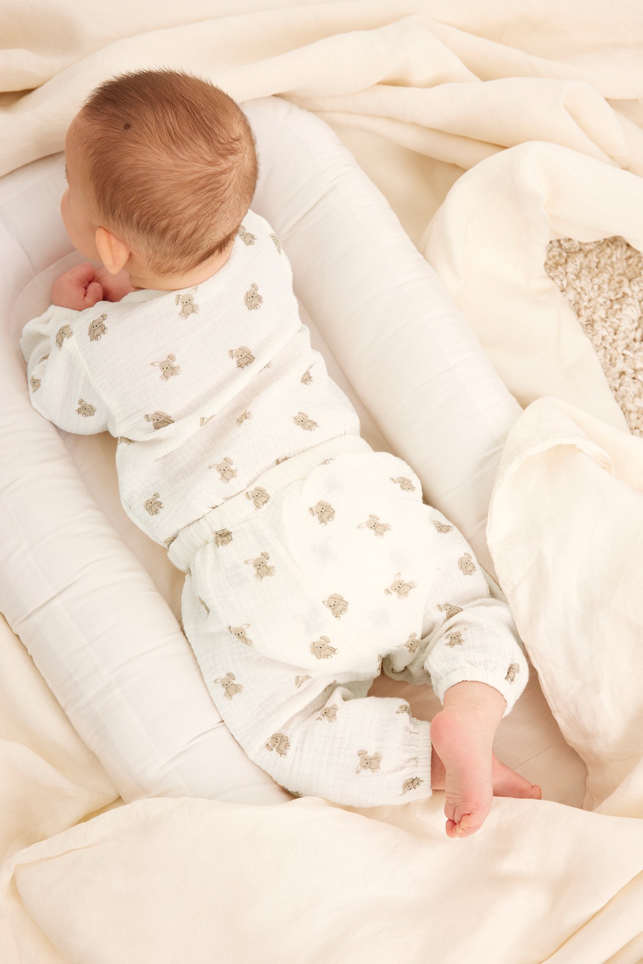 Ylhäältä takaa kuvattu vauva yllään luonnonvalkoinen kuviollinen puuvillamusliininen pitkähihainen kietaisubody ja yhteensopivat housut.