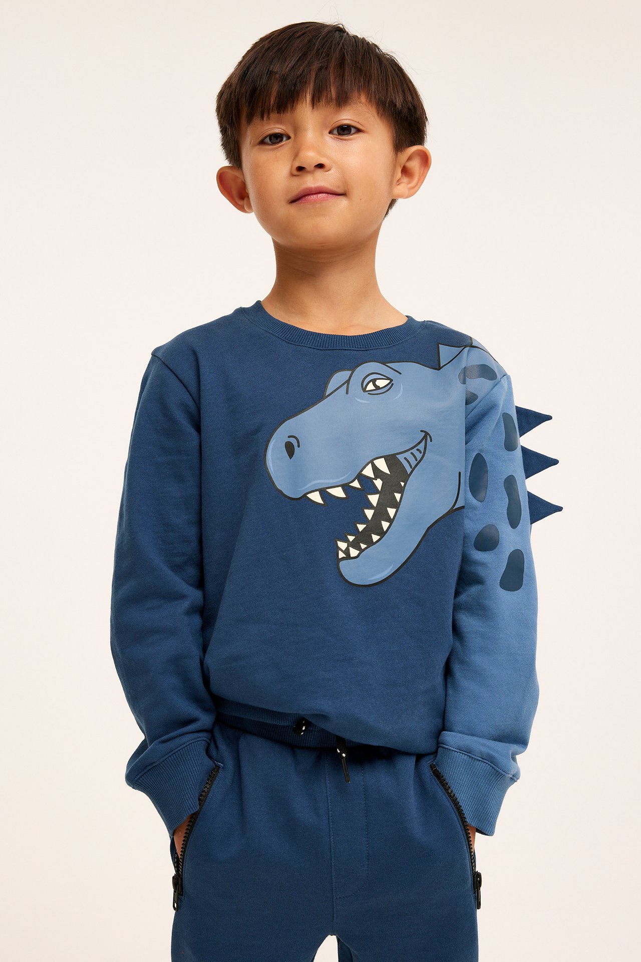 Edestäpäin, lapsi yllään tummansininen puuvillainen dinosauruskuvioinen collegepaita ja yhteensopivat collegehousut.