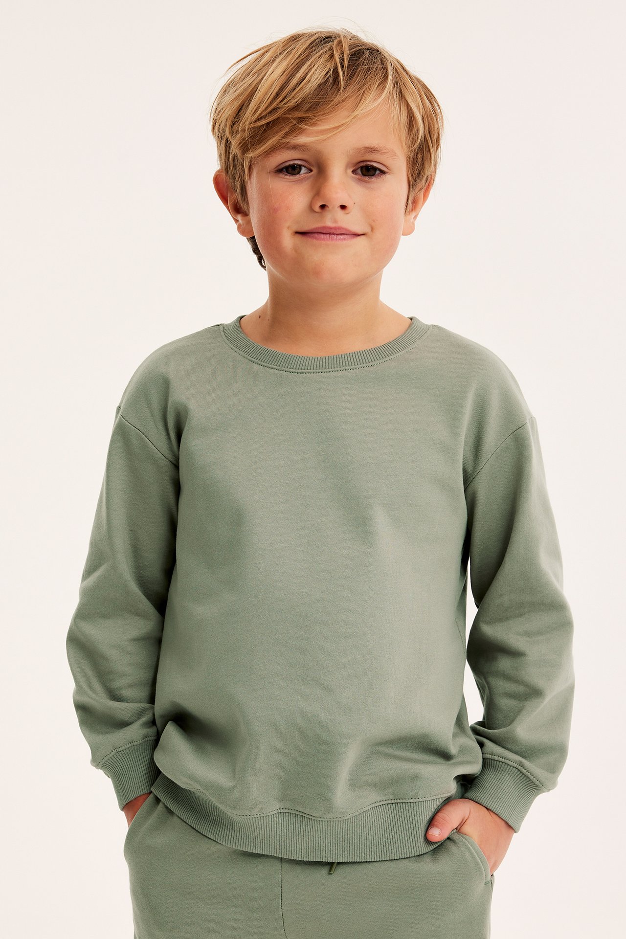 Frontvy av ett barn i en enfärgad grön bomullssweatshirt med ribbade detaljer och matchande sweatpants.