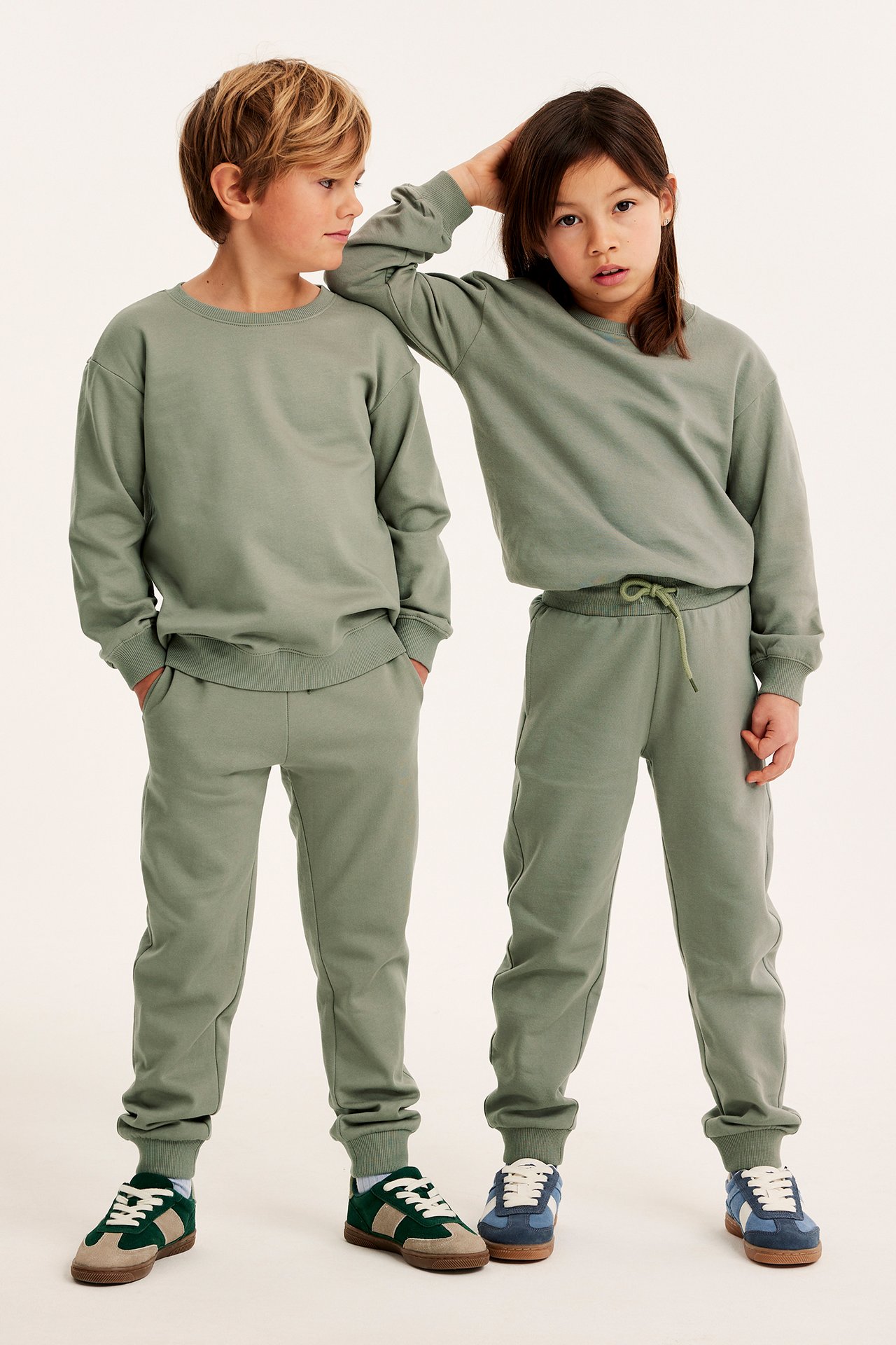 Framifrån: Pojke och flicka i gröna bomullssweatshirts med ribbade muddar, matchande sweatpants och sneakers.