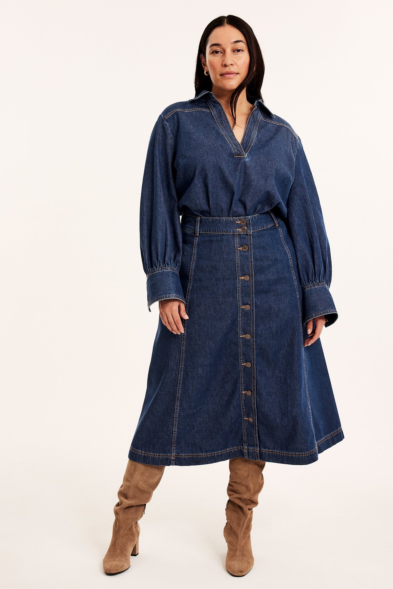 Widok z przodu kobiety w ciemnoniebieskiej rozkloszowanej spódnicy jeansowej na guziki, pasującej koszuli i brązowych zamszowych kozakach.