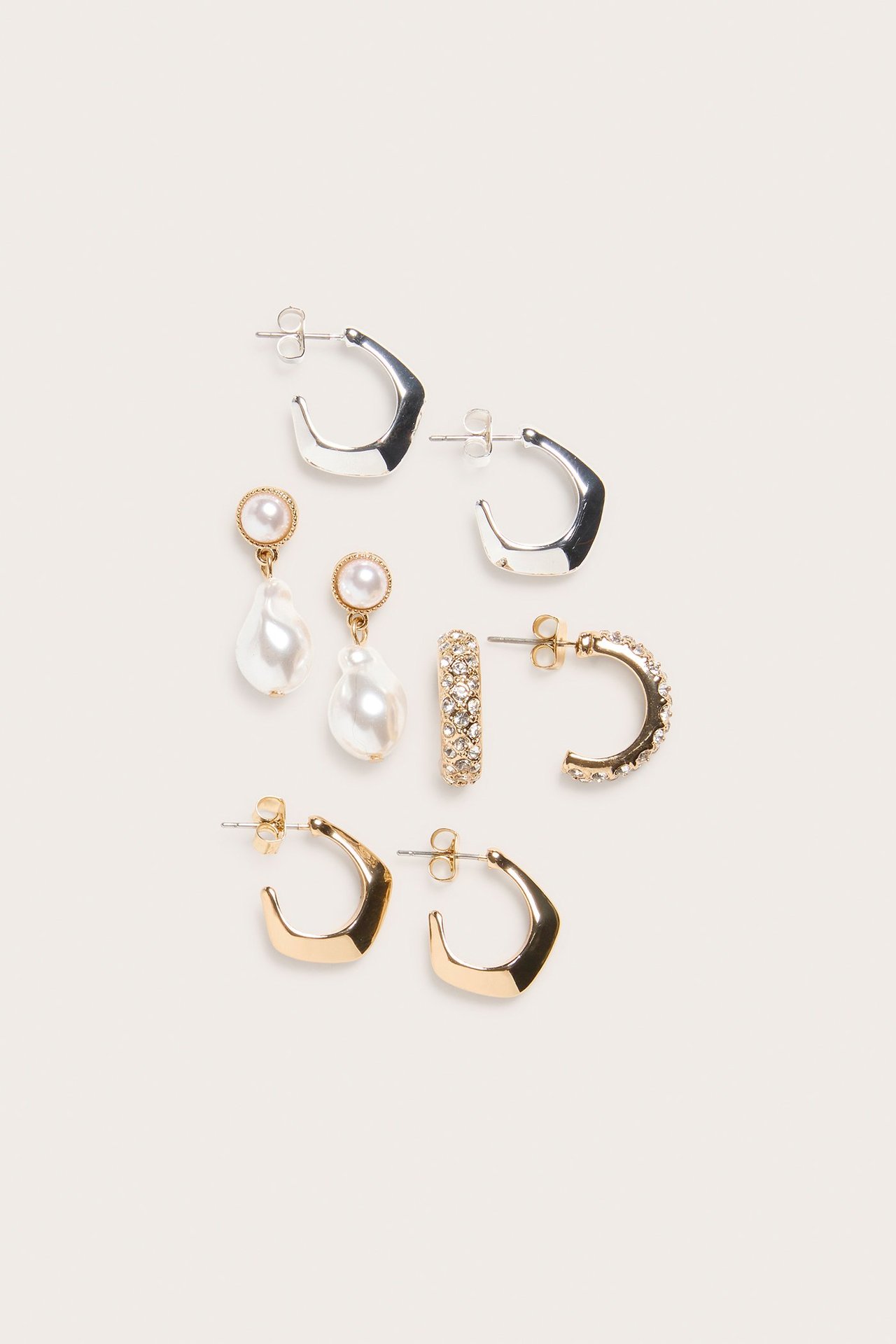 Edestä näkymä 4-pakkauksesta korvakoruja: kultaiset ja hopeiset kulmikkaat renkaat, kultaiset helmipisarat, ja kultaiset strassipuolirenkaat.