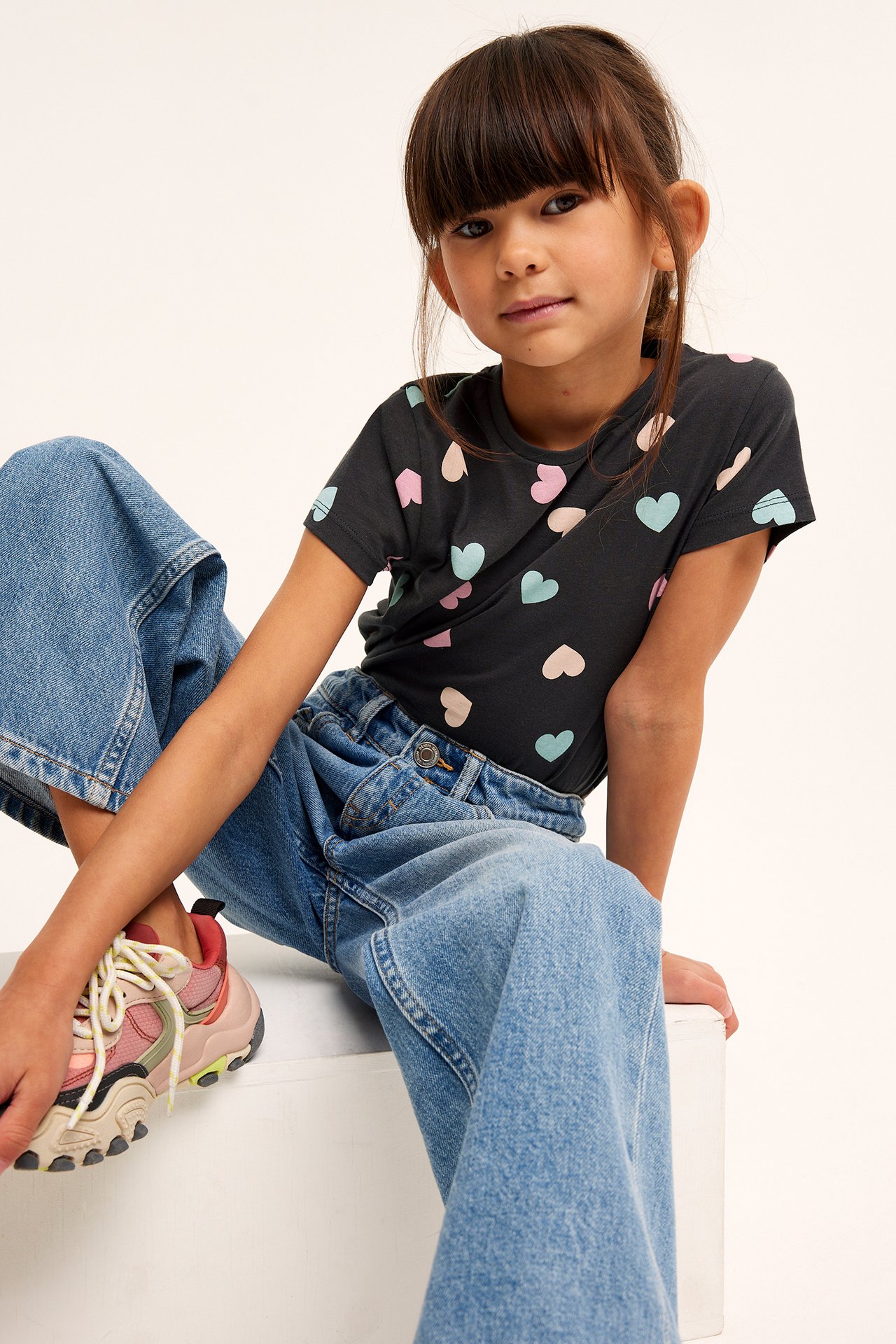 Dziewczynka w czarnym t-shircie z serduszkami, niebieskie jeansy, buty sportowe, front.
