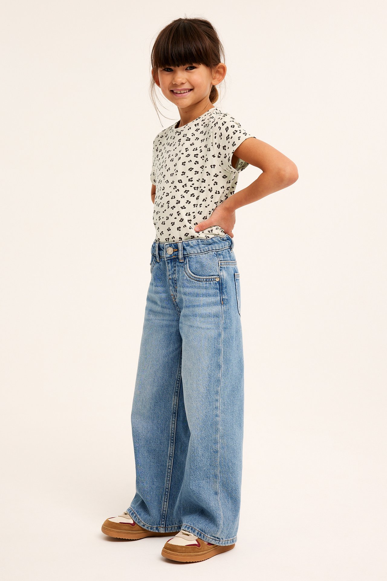 Frontvy av ett barn i beige leopardmönstrad bomulls-t-shirt, vida blå jeans och bruna sneakers.