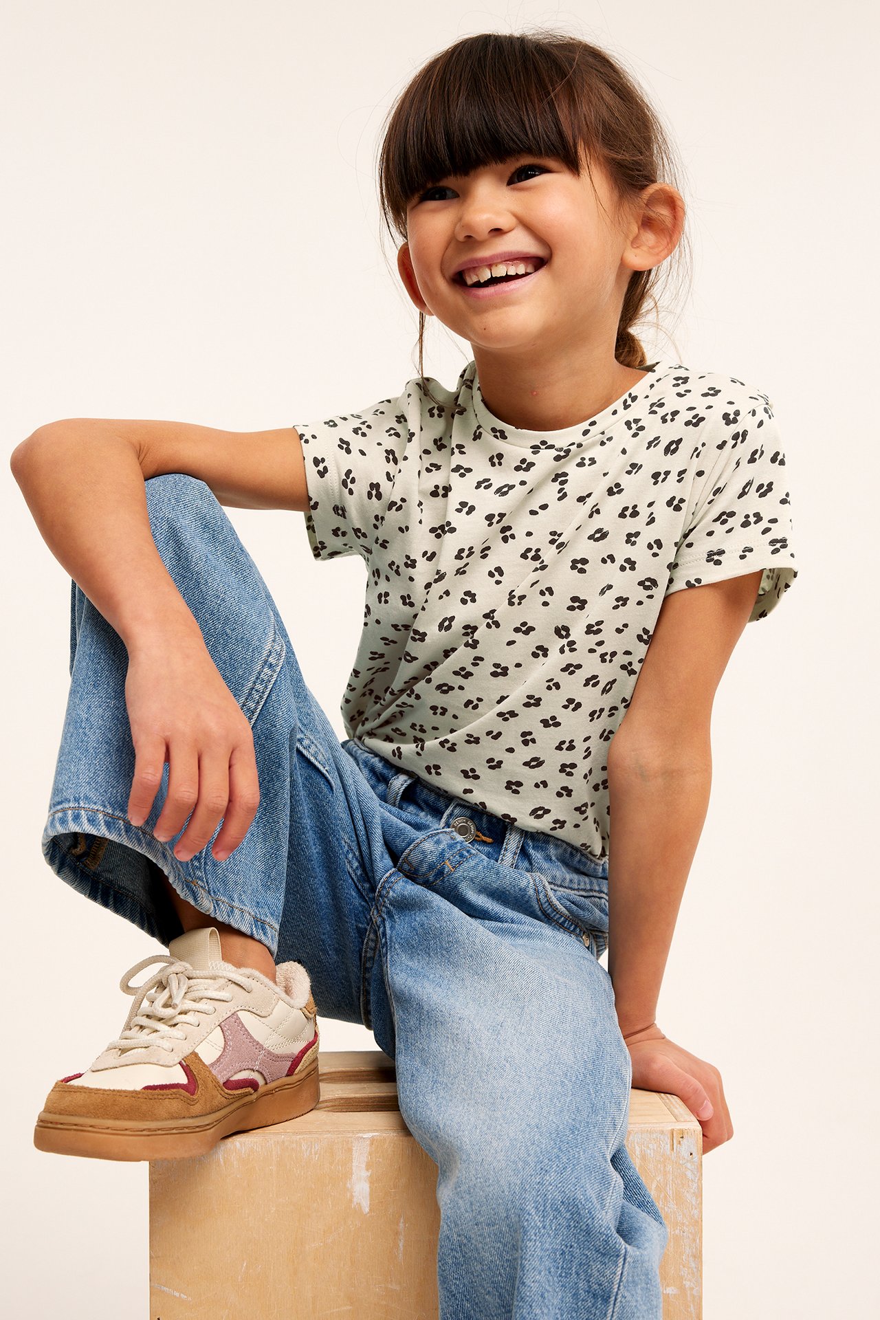 Framifrån, leende barn i ljusgrön leopardmönstrad t-shirt, blå jeans och flerfärgade sneakers.