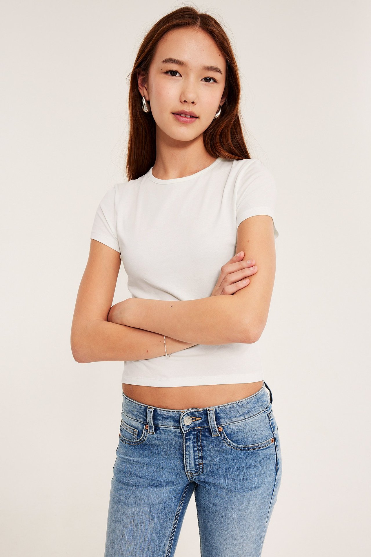 Edestäpäin, lapsi luonnonvalkoisessa istuvassa lyhythihaisessa T-paidassa, sinisissä farkuissa, hopeakorvakoruissa ja ohuessa hopearannekorussa.