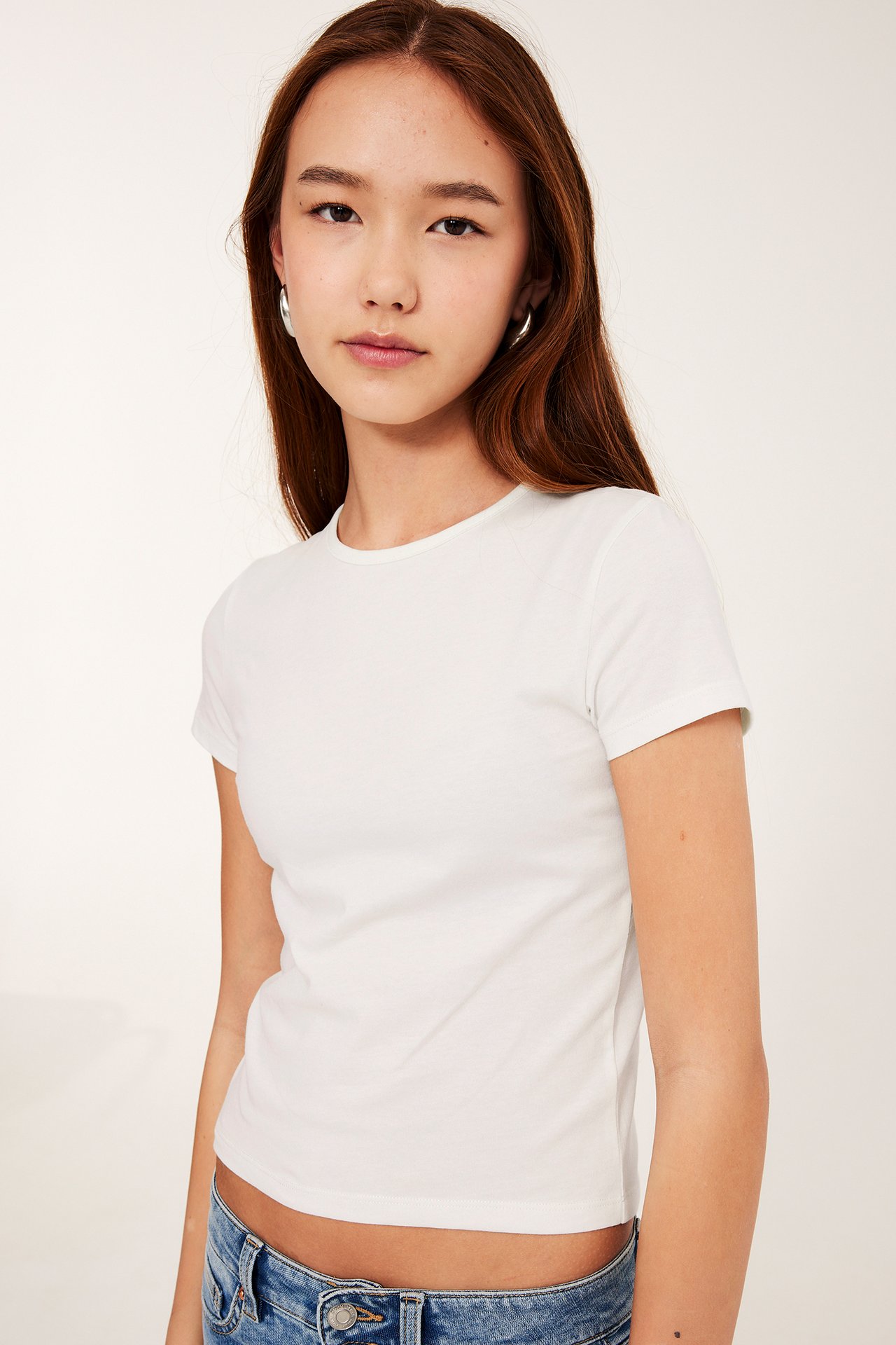 Framifrån: kvinna i offwhite, kortärmad, slim-fit t-shirt, blå jeans och silverfärgade ringörhängen.