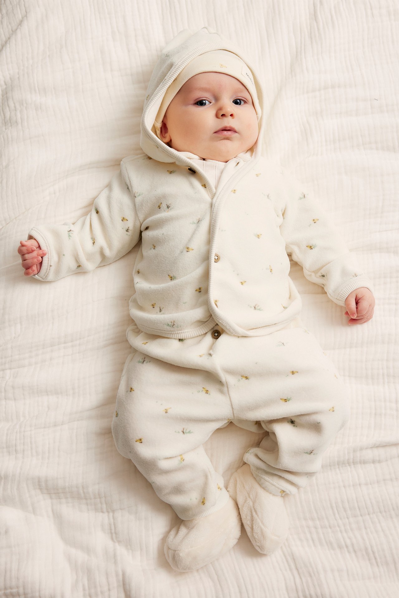 Ovenfra: Baby i offwhite mønstret velourhettegenser med knapper, matchende bukser, lue og tøfler.